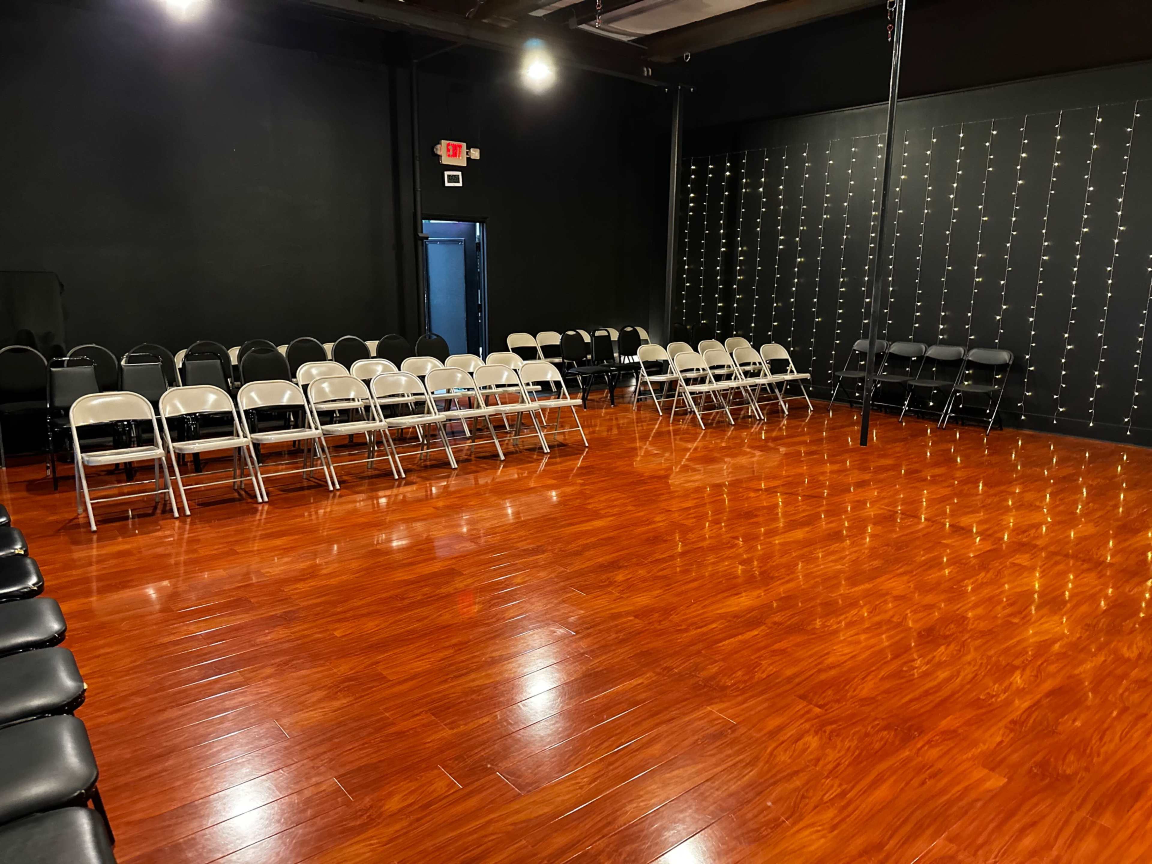 The image shows a rehearsal or performance space with rows of folding chairs set up on a polished wooden floor, illuminated by string lights on a black wall.