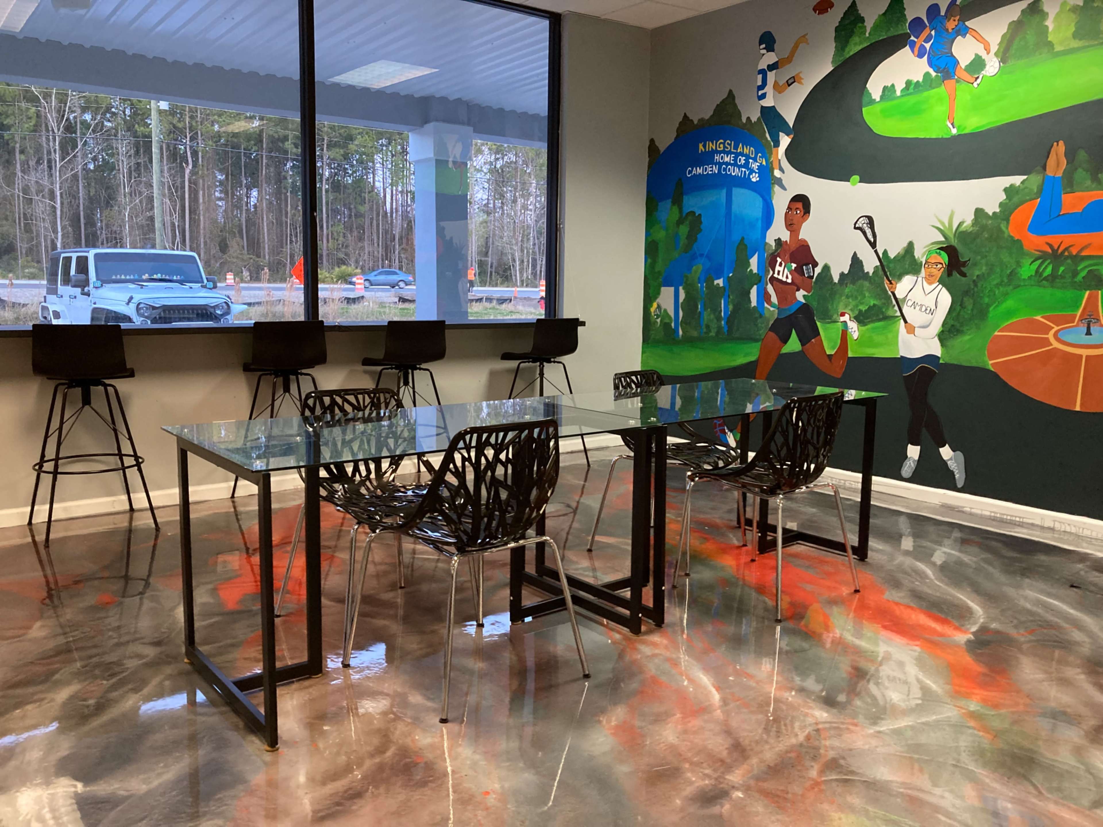 A modern workspace features a glass table surrounded by black chairs and a colorful mural on the wall.