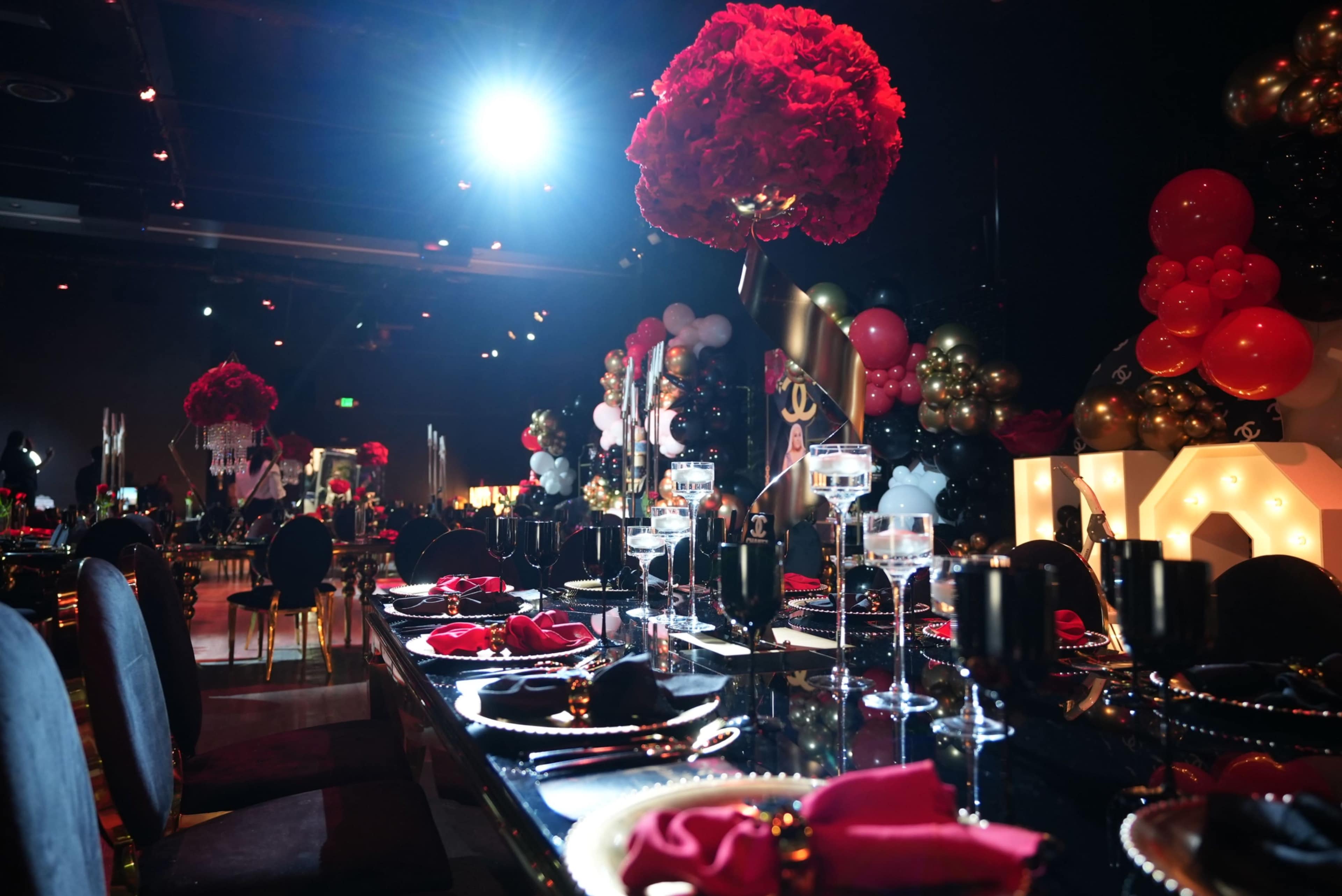 The image shows an elegantly decorated dining space with a long table set with black and gold tableware, red napkins, and tall floral centerpieces.
