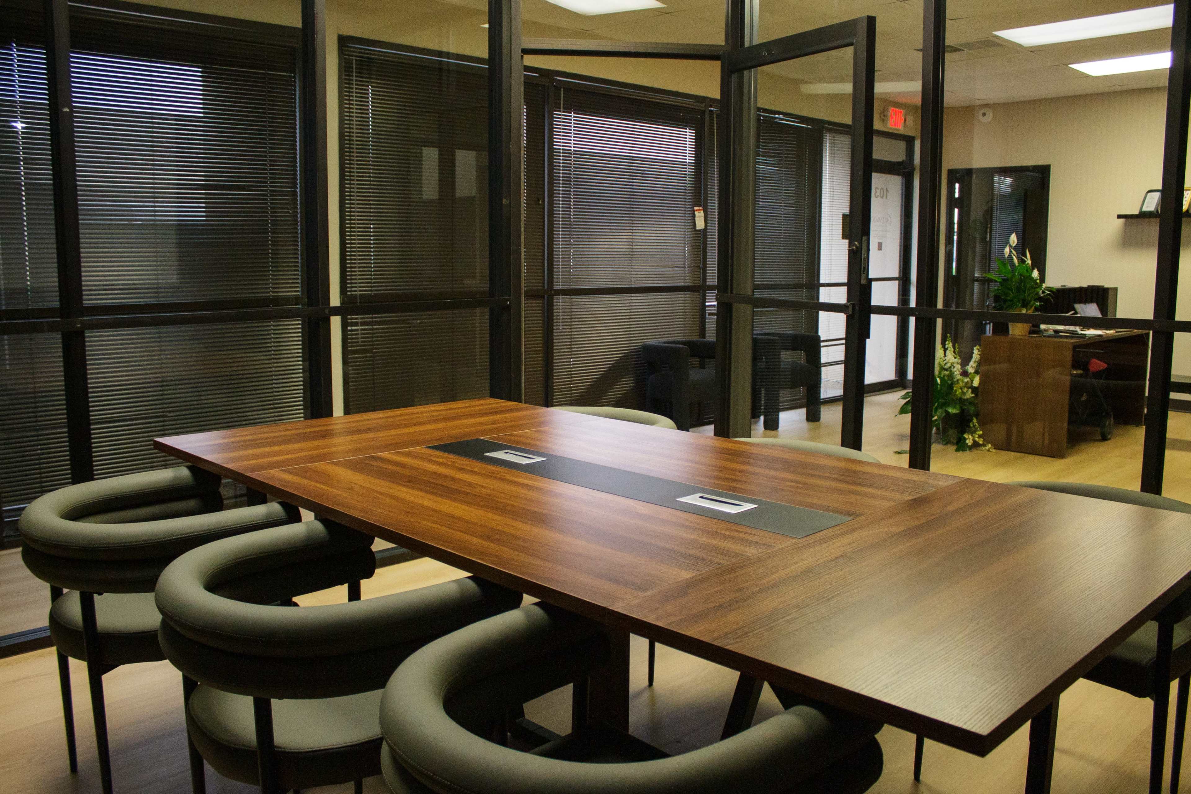 The image shows a modern conference room with a wooden table surrounded by black, rounded chairs, enclosed by glass walls.