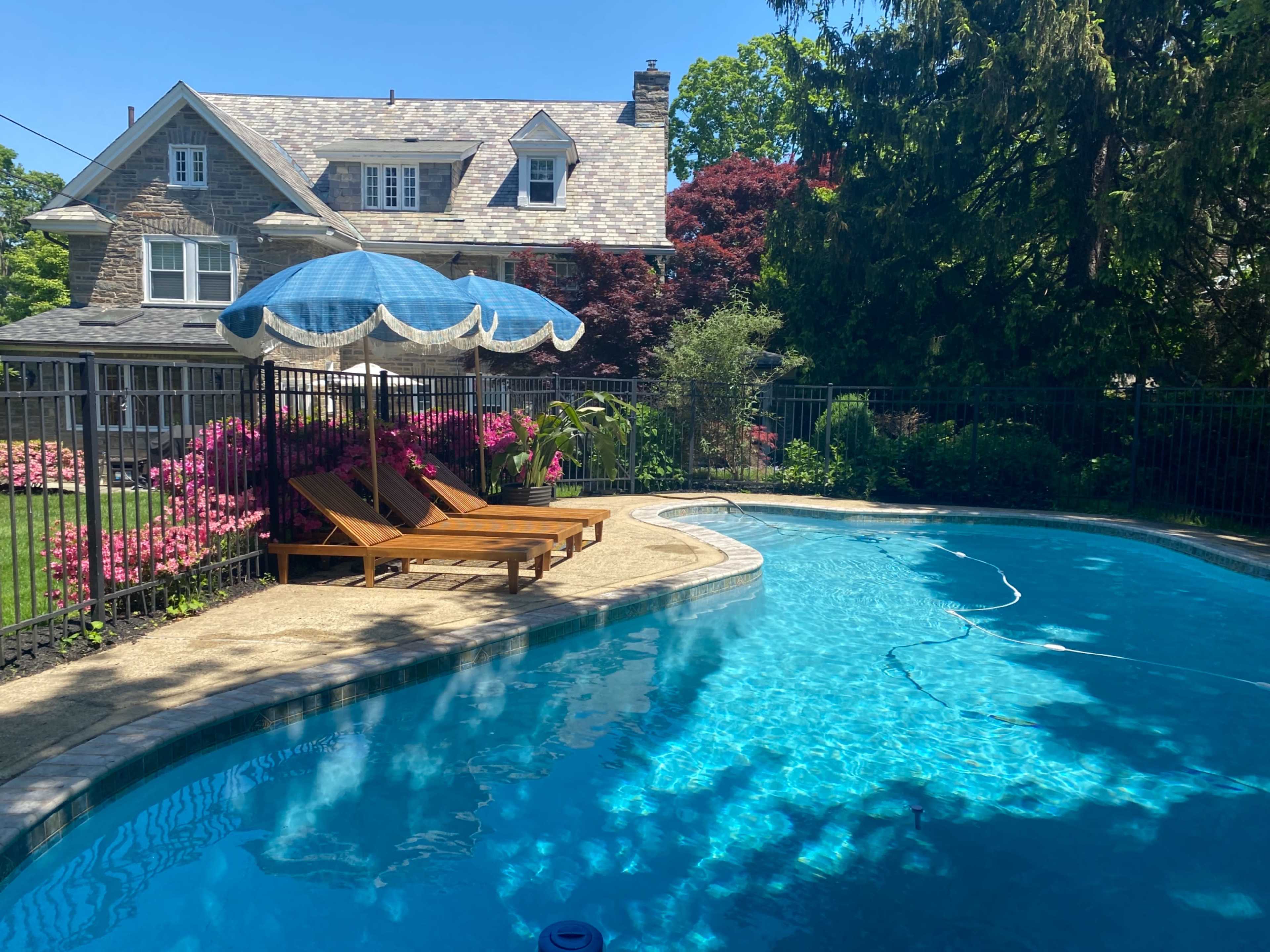 A swimming pool with clear blue water is surrounded by a fence, deck chairs, and vibrant flowers beneath a blue umbrella.