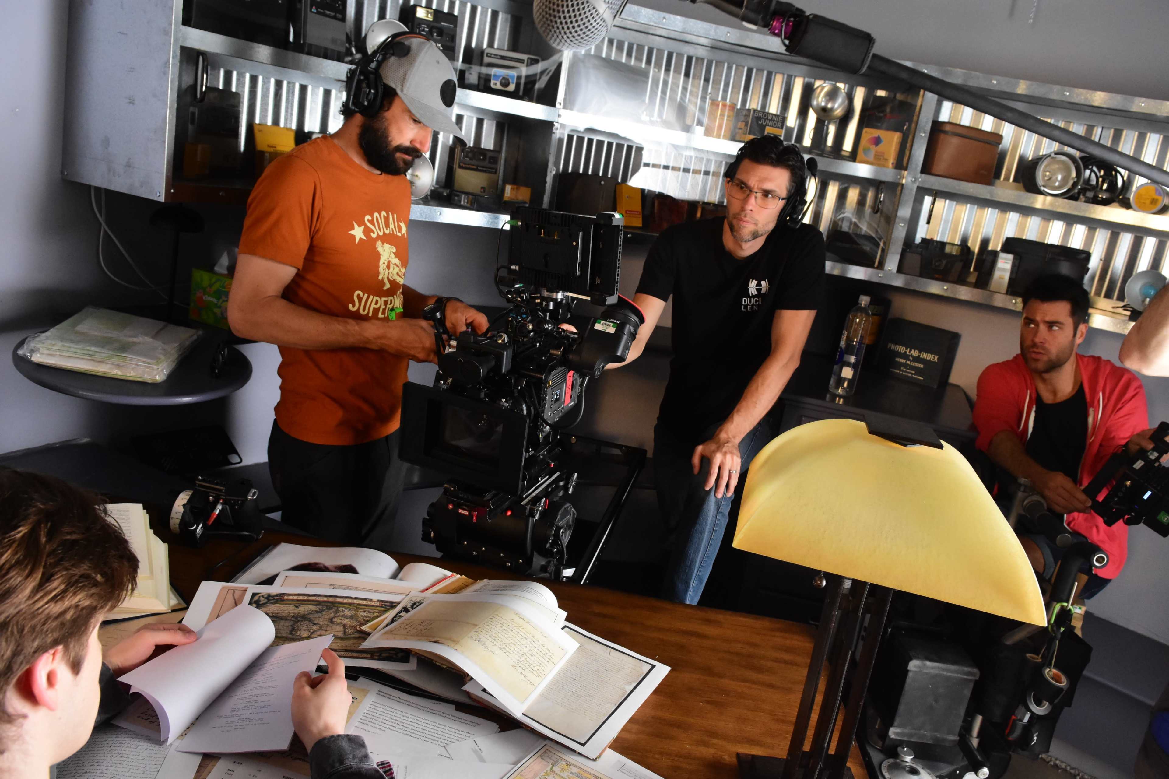 A crew of filmmakers is gathered in a dimly lit room, working with camera equipment and scripts on a wooden table.