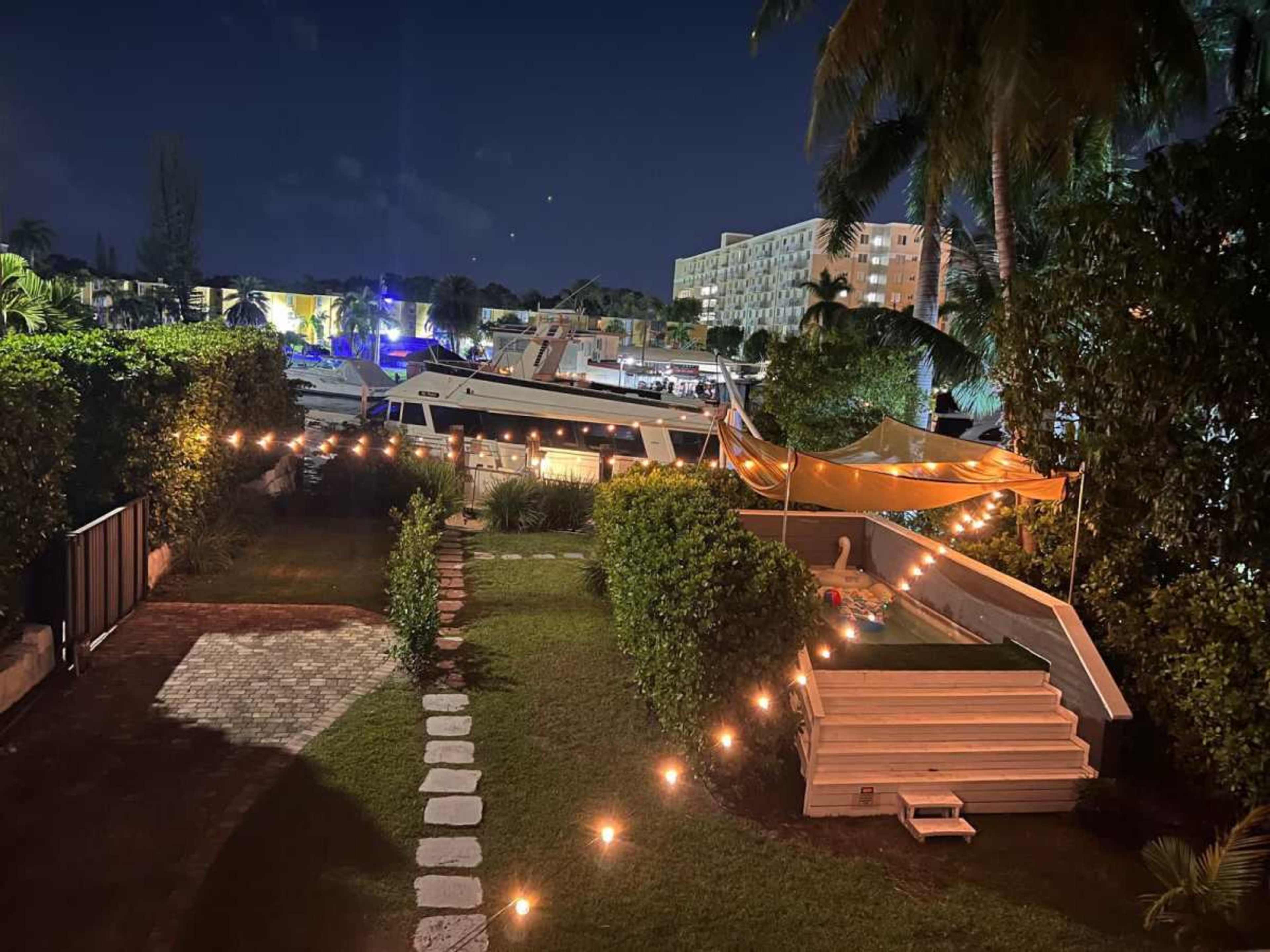 A well-lit backyard features a pathway of stones leading through greenery, with a boat moored nearby and string lights illuminating a seating area under a tent.
