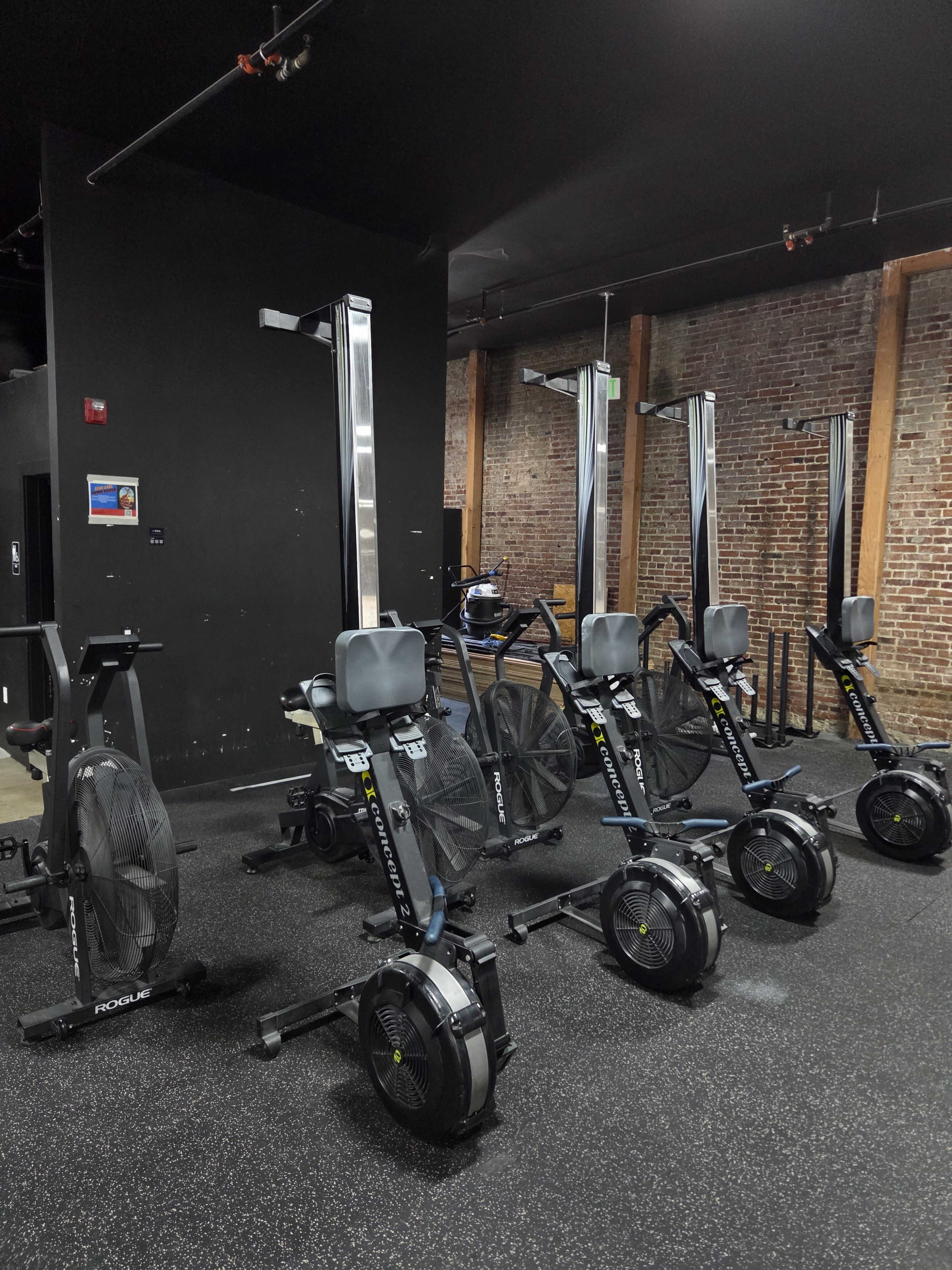 The image shows a gym area featuring several rowing machines arranged in a row against a backdrop of exposed brick walls.