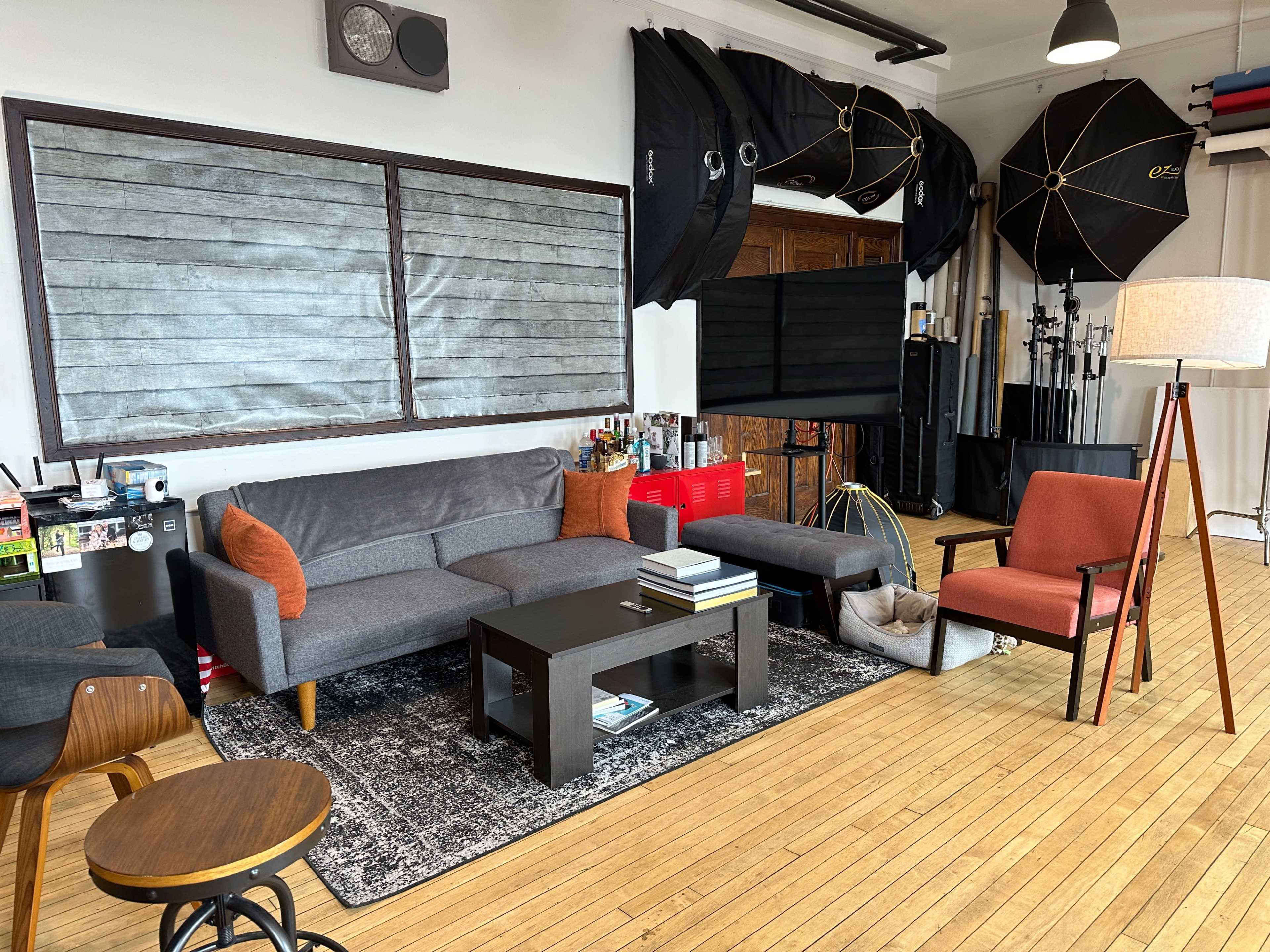 A modern studio space features a gray sofa, a black coffee table, an orange-accented chair, and various lighting equipment in the background.