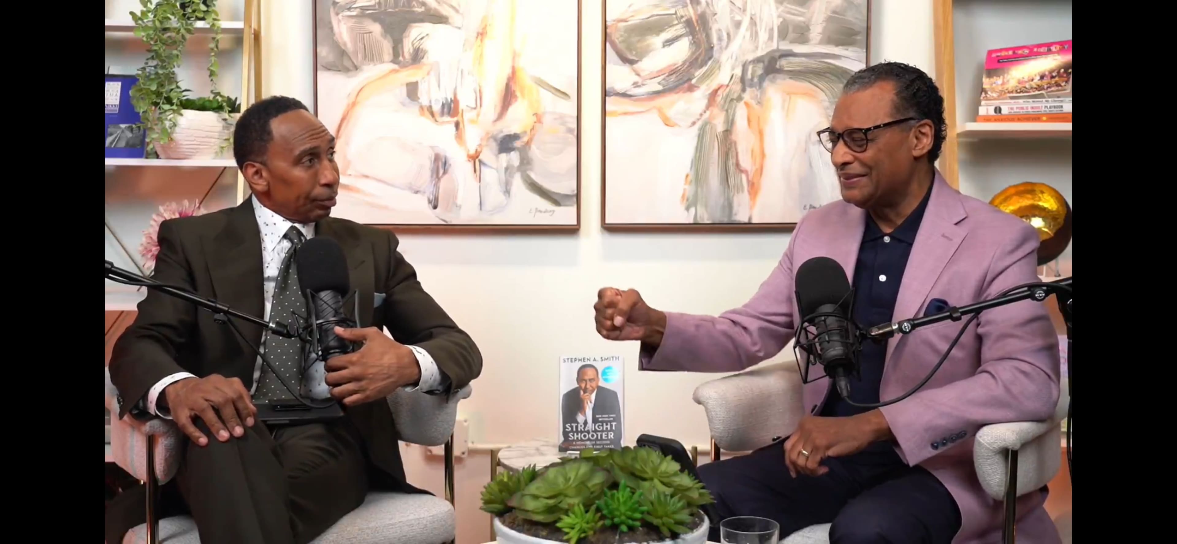 Two men are seated in modern chairs with microphones in front of them, discussing a book titled "Straight Shooter" on a table adorned with a succulent plant.