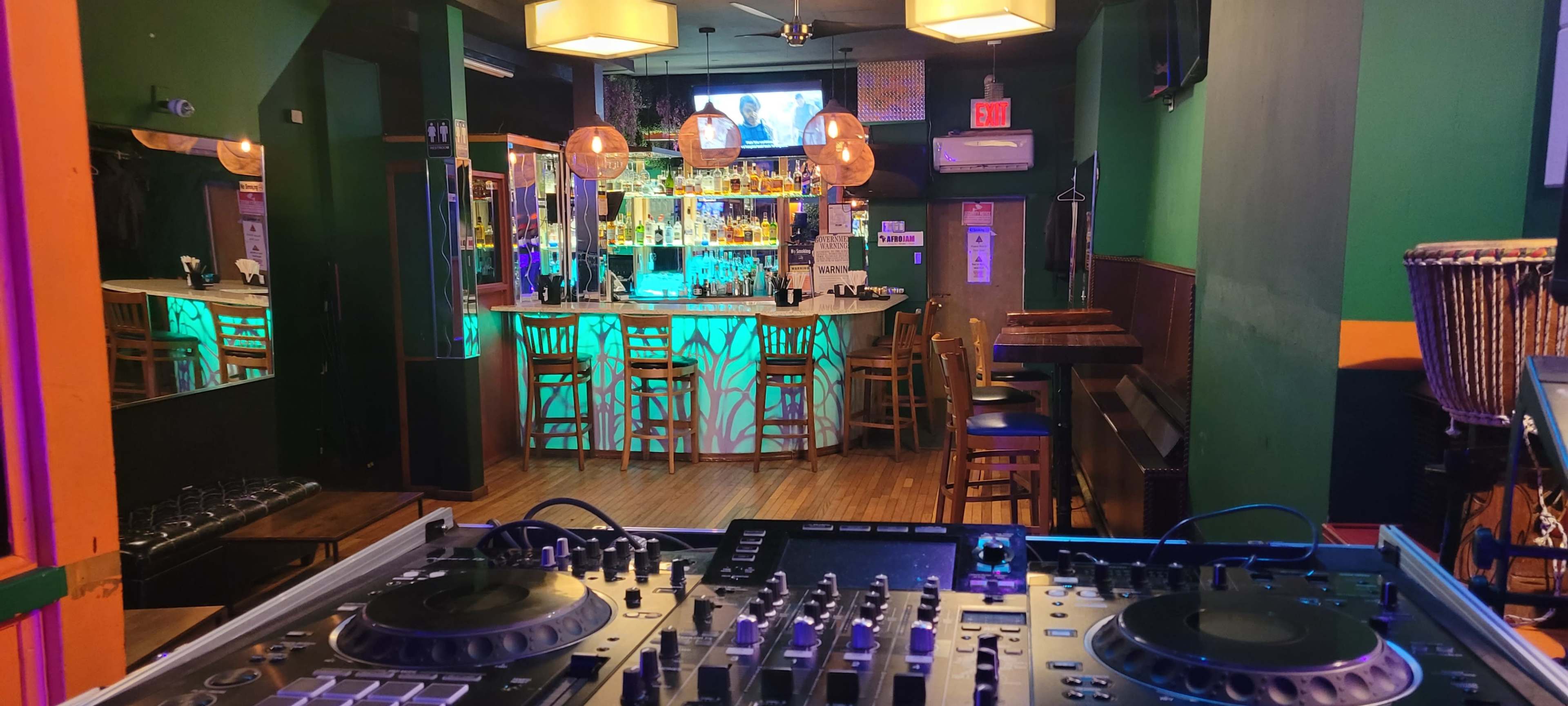 A DJ setup with turntables is positioned in the foreground, overlooking a brightly lit bar area filled with stools and shelves lined with various bottles.