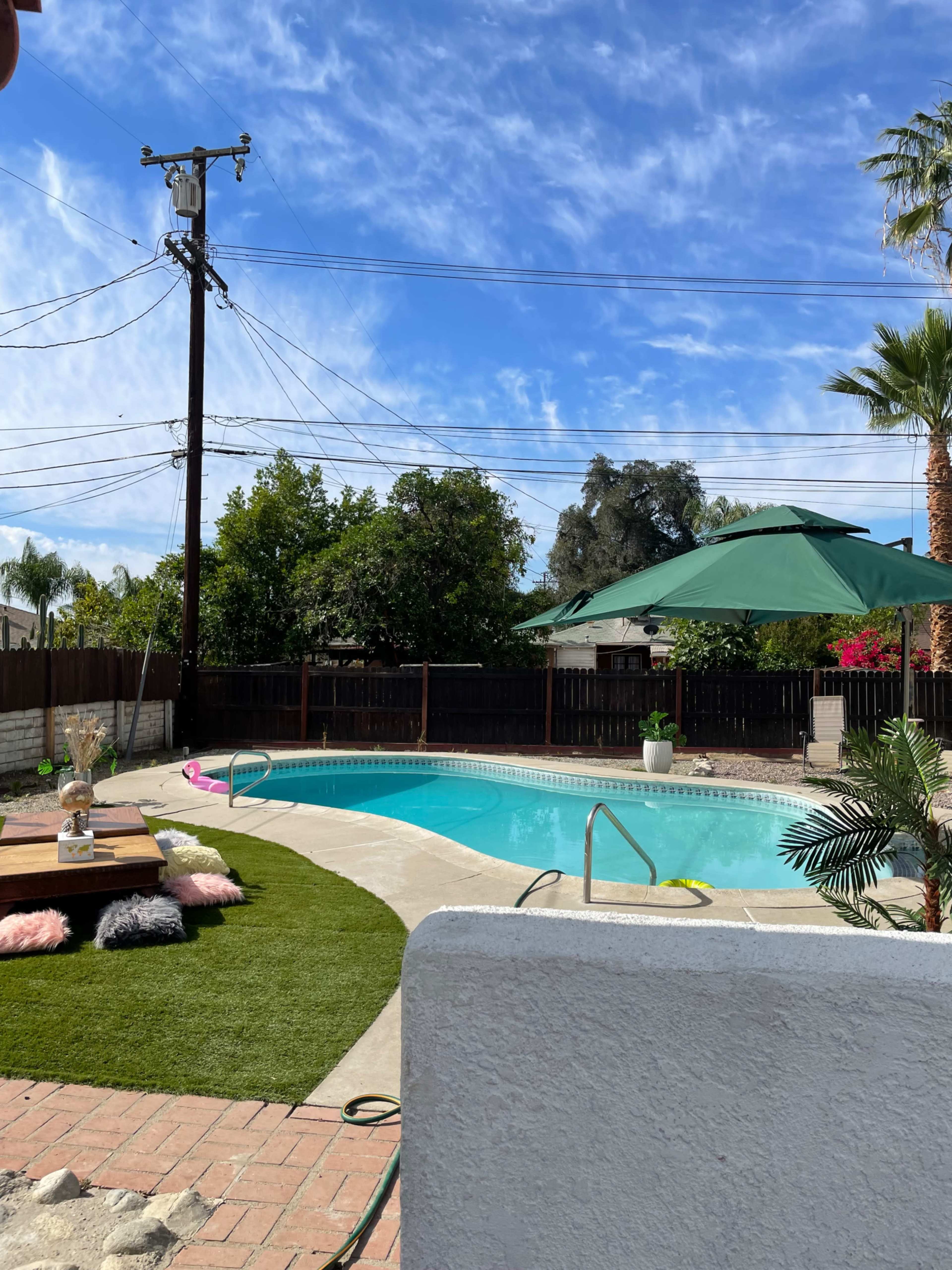 The image shows a swimming pool surrounded by green grass, a wooden deck with seating, and palm trees, under a clear blue sky with power lines in the background.