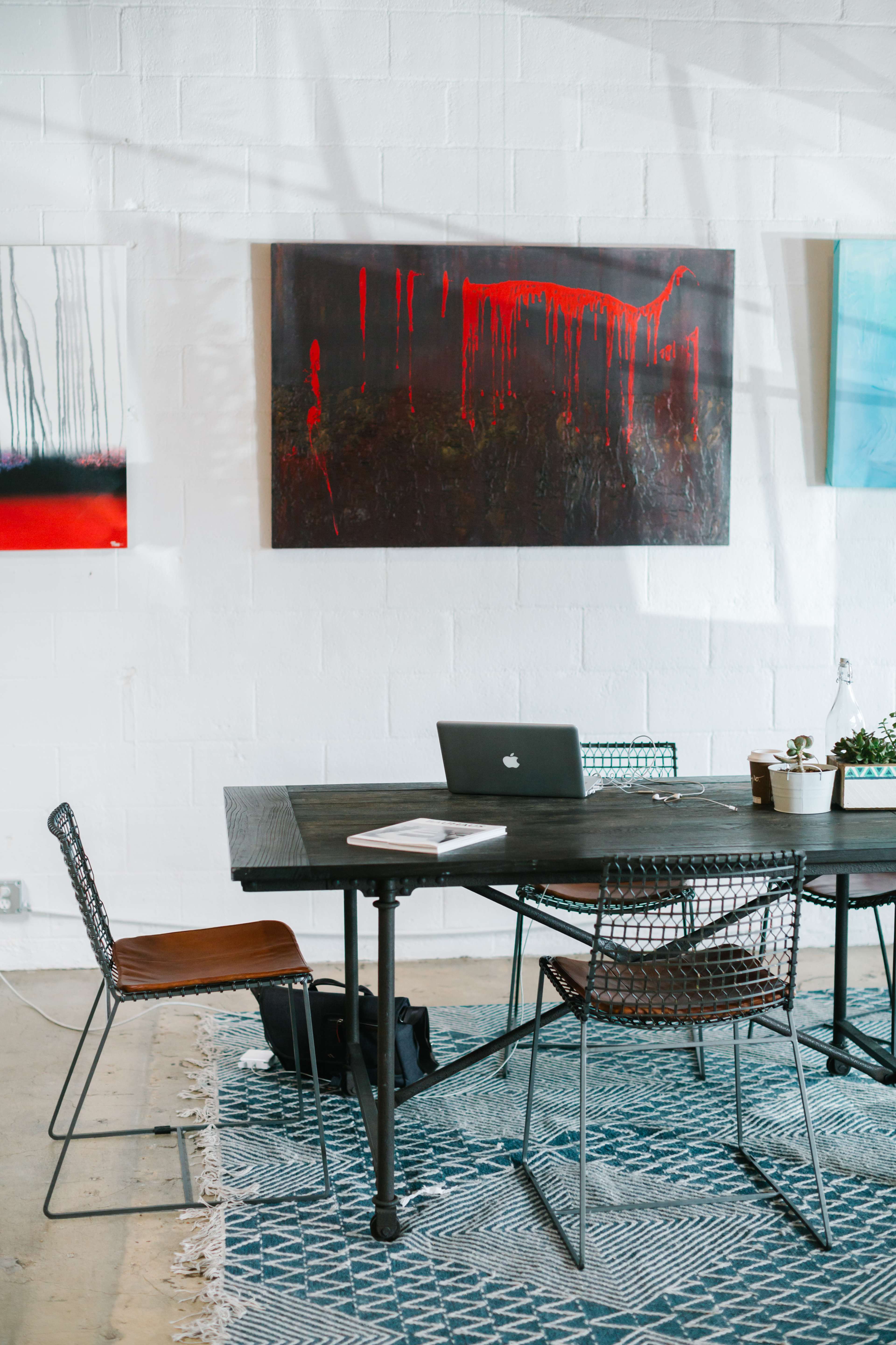 A workspace features a dark wooden table with a laptop, surrounded by wire chairs, against a backdrop of white walls adorned with abstract paintings.