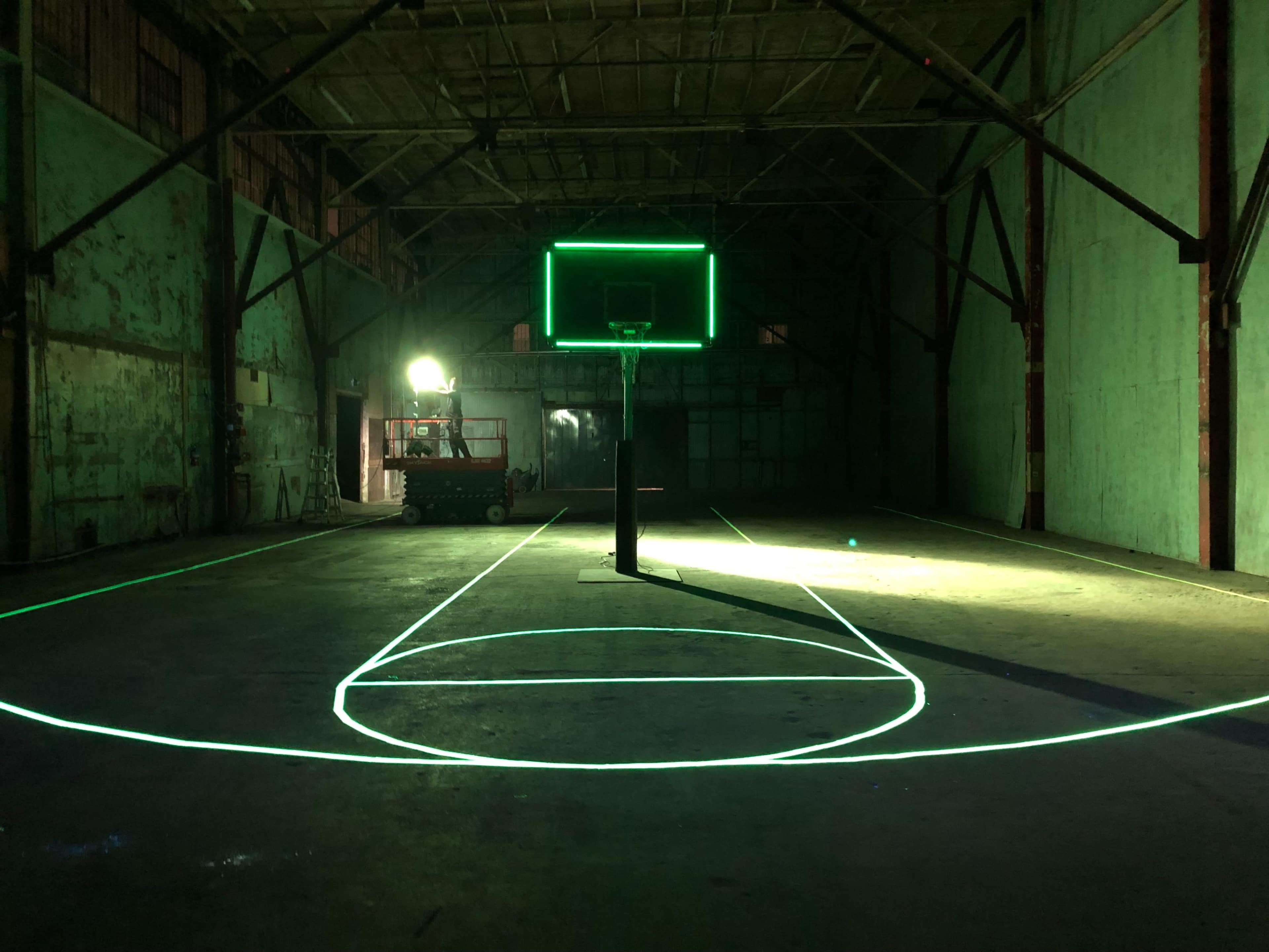 A dimly lit basketball court is outlined with green light, featuring a hoop and backboard in an industrial warehouse.