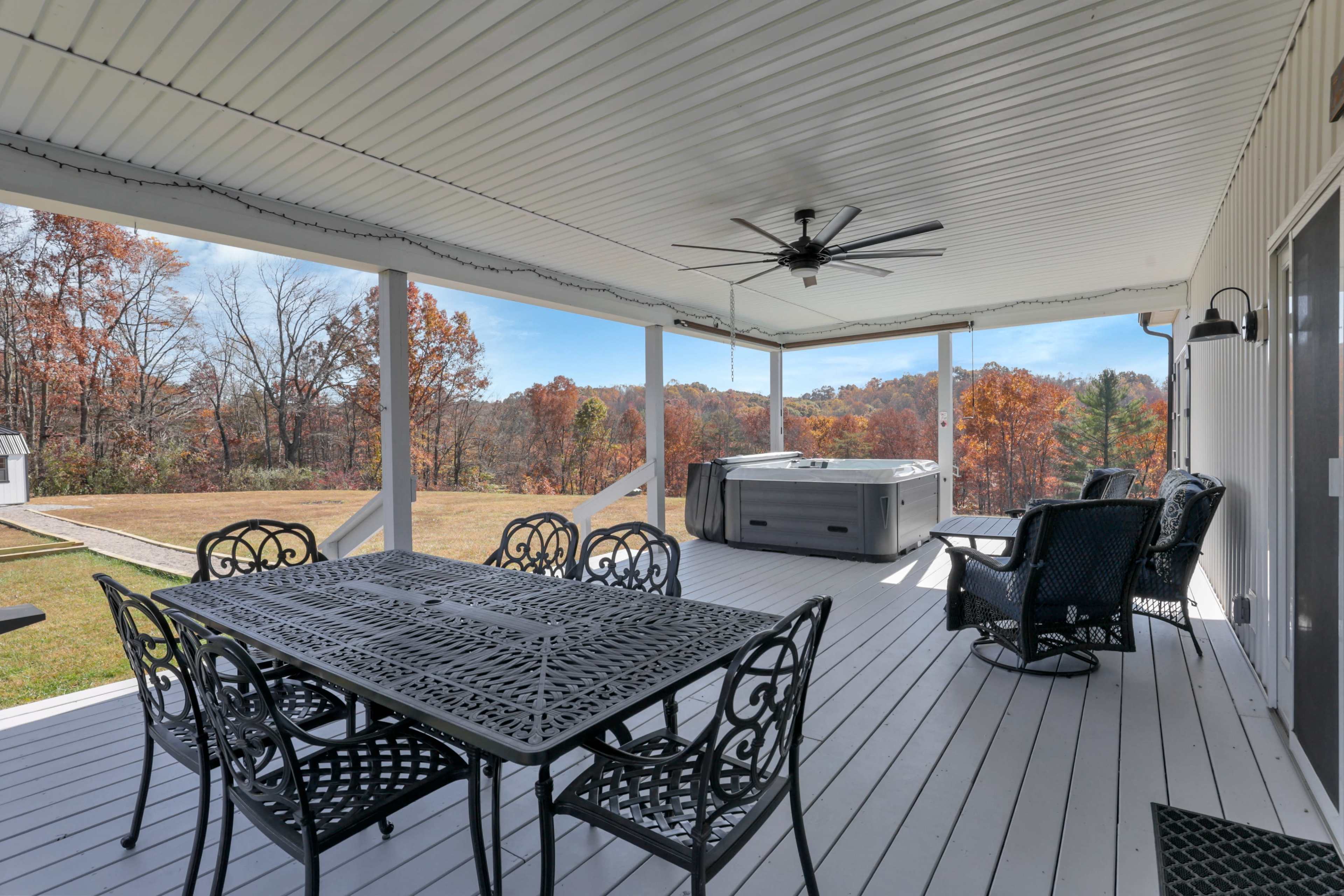 A covered porch features a dining table, several chairs, and a hot tub, overlooking a wooded landscape with autumn foliage.