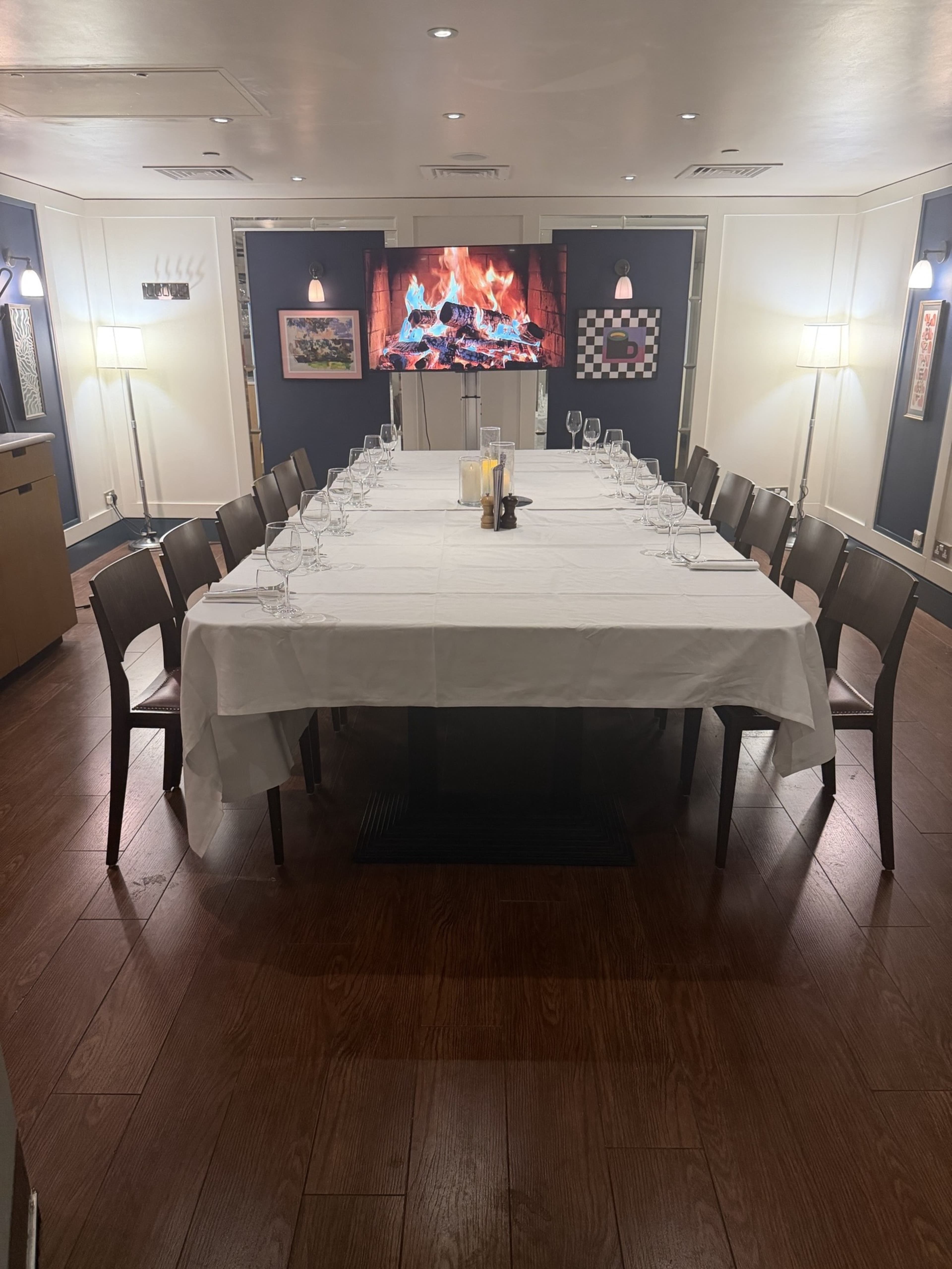 A long table set for a formal dinner is placed in a well-lit private dining room, with a fireplace display on the wall and framed artwork around the space.