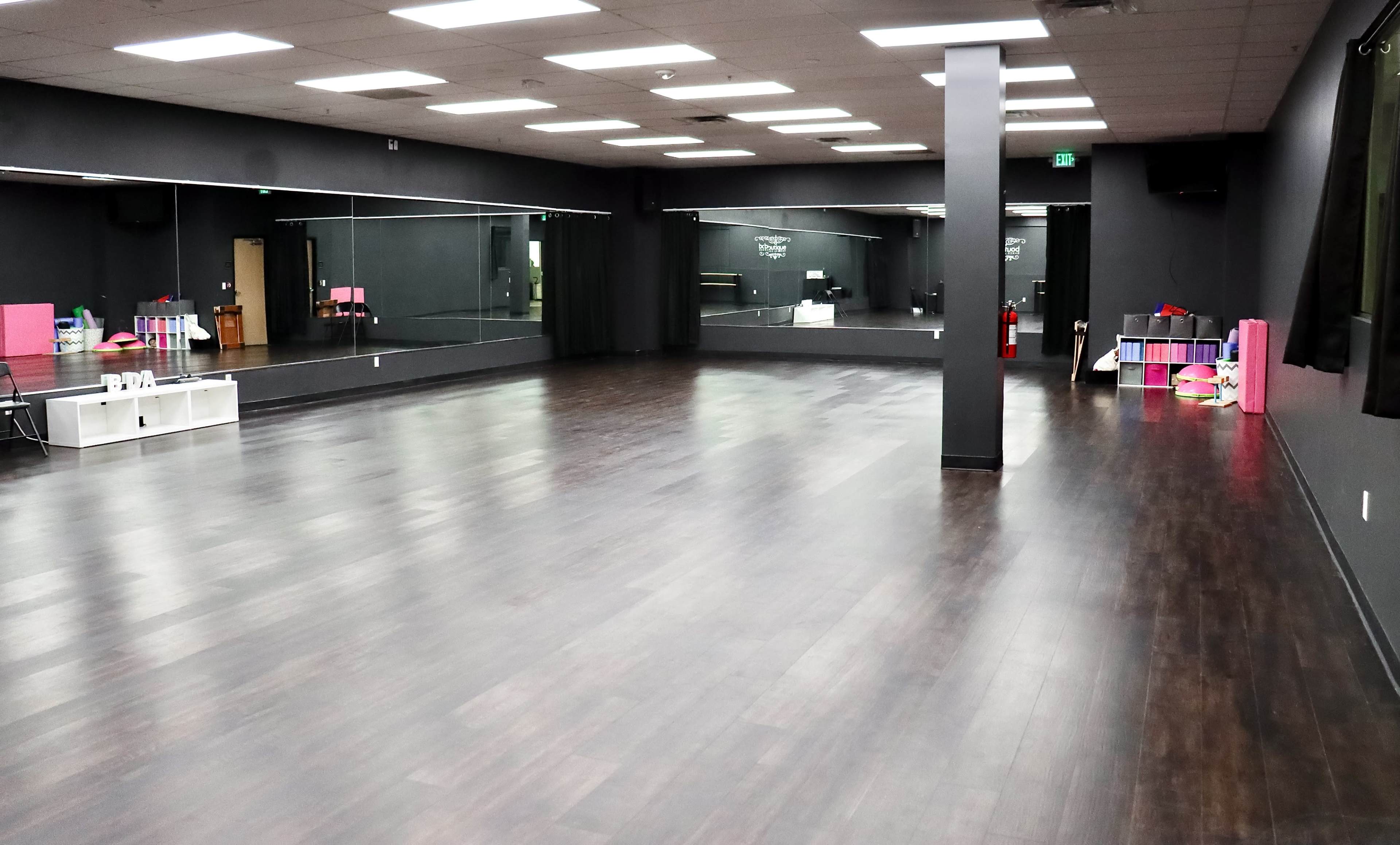 A spacious dance studio with dark wood flooring, black walls, and large mirrors on one side.