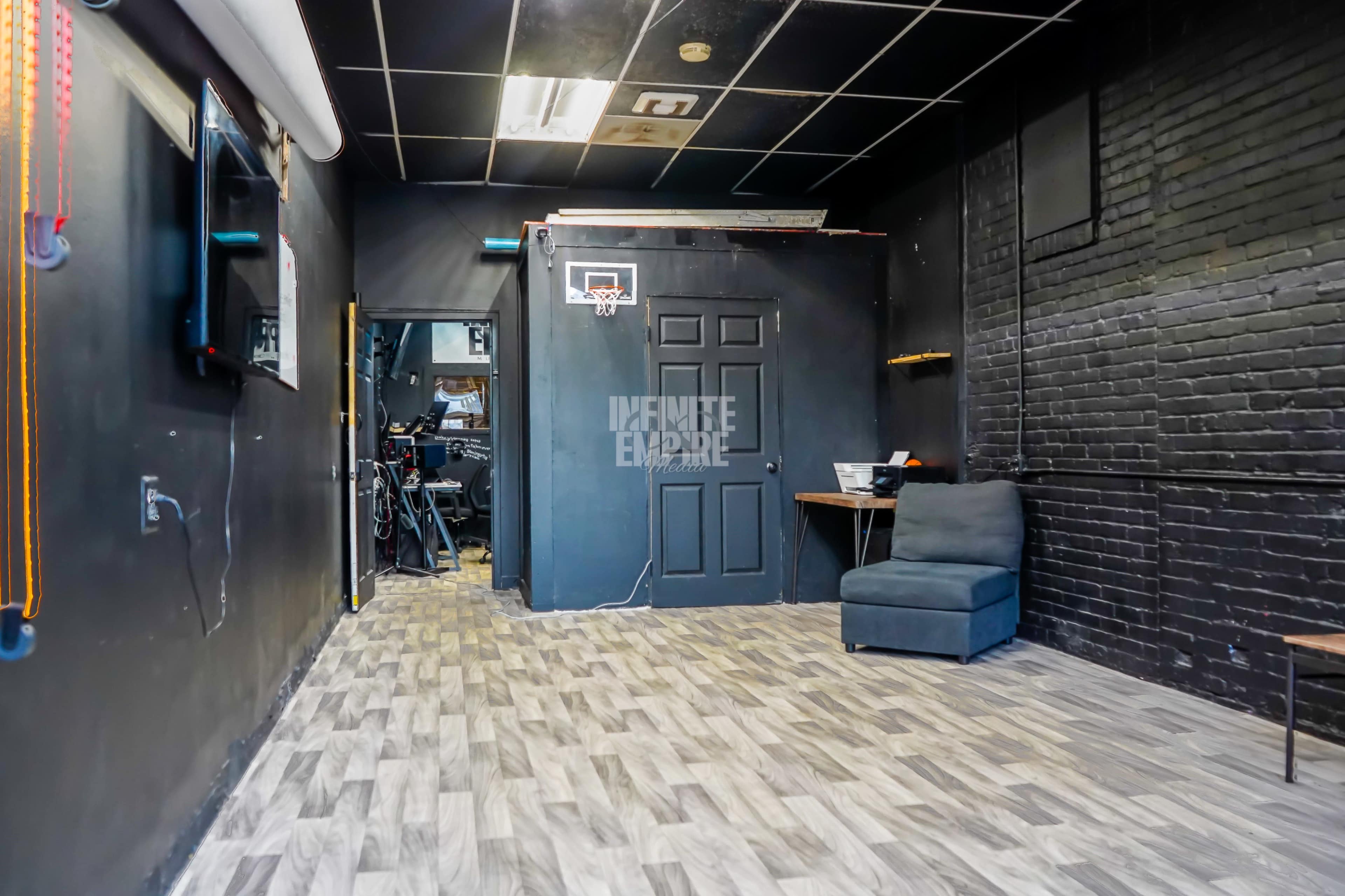 The image shows a dimly lit room with dark walls, a small basketball hoop mounted on the door, and a blue chair in the corner, along with a desk in the background.