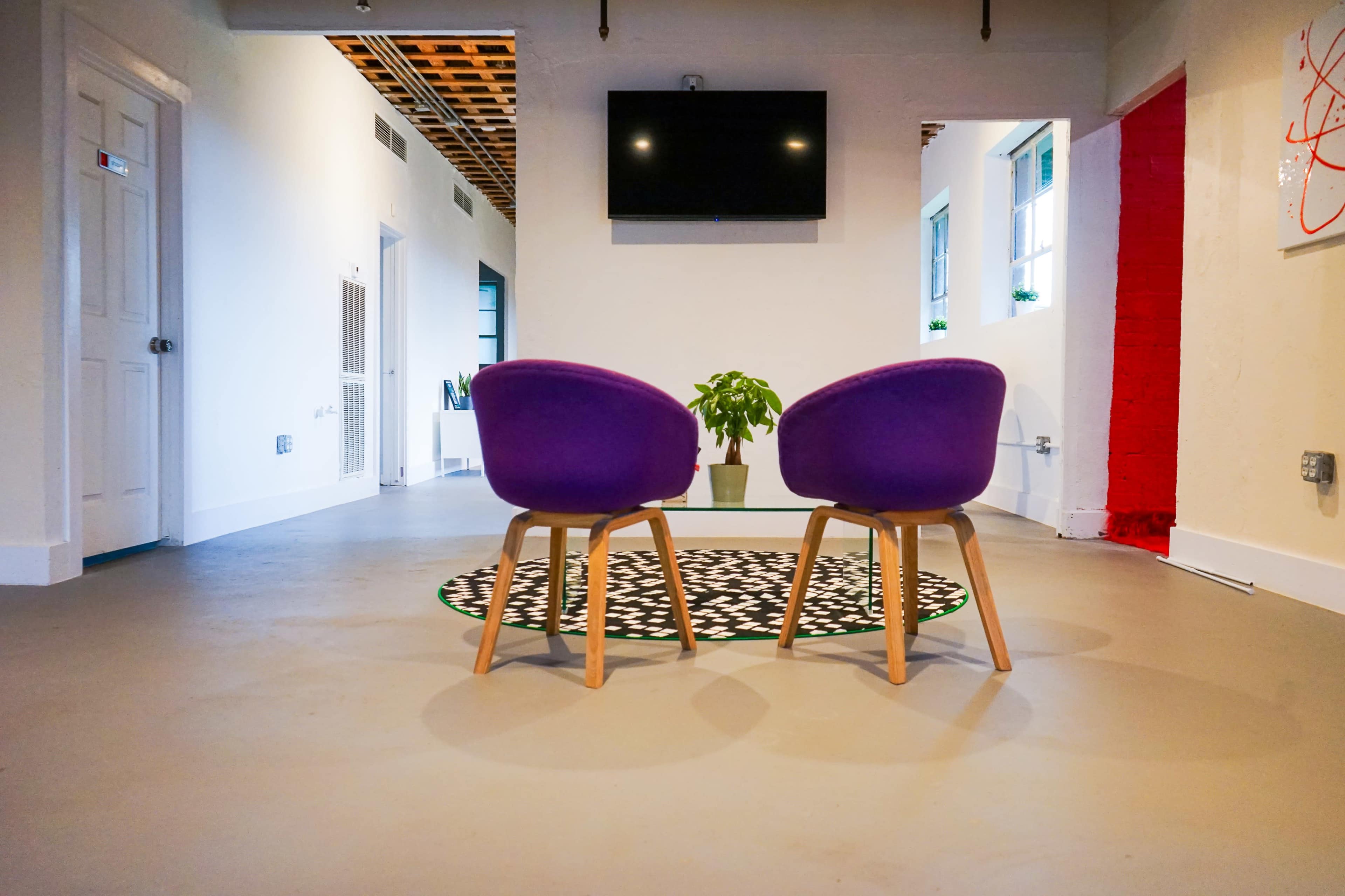 Two purple chairs face each other on a patterned rug in a well-lit room with white walls and a television mounted on the wall.