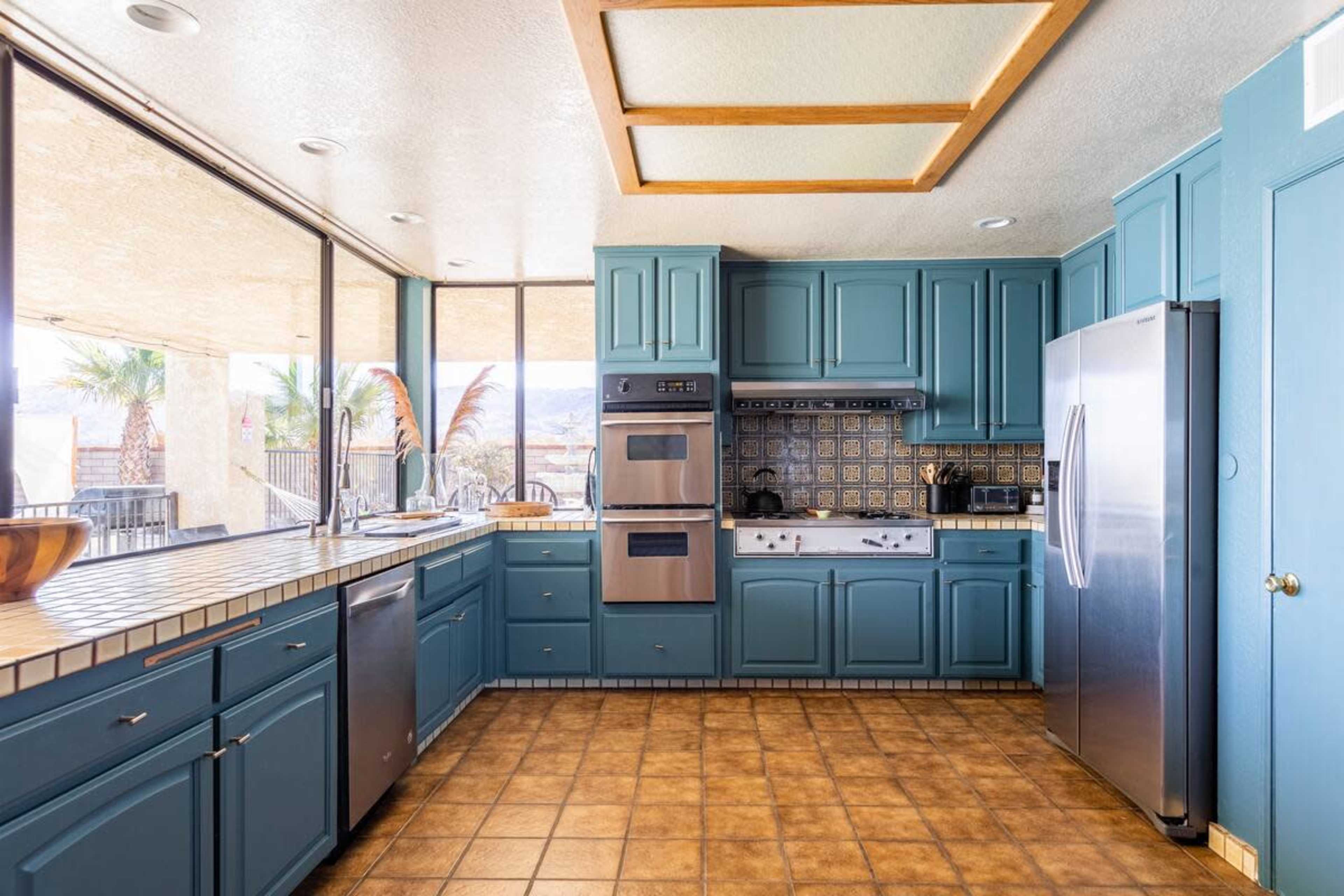 The image shows a kitchen featuring blue cabinetry, stainless steel appliances, and large windows that provide natural light and views of the outside.