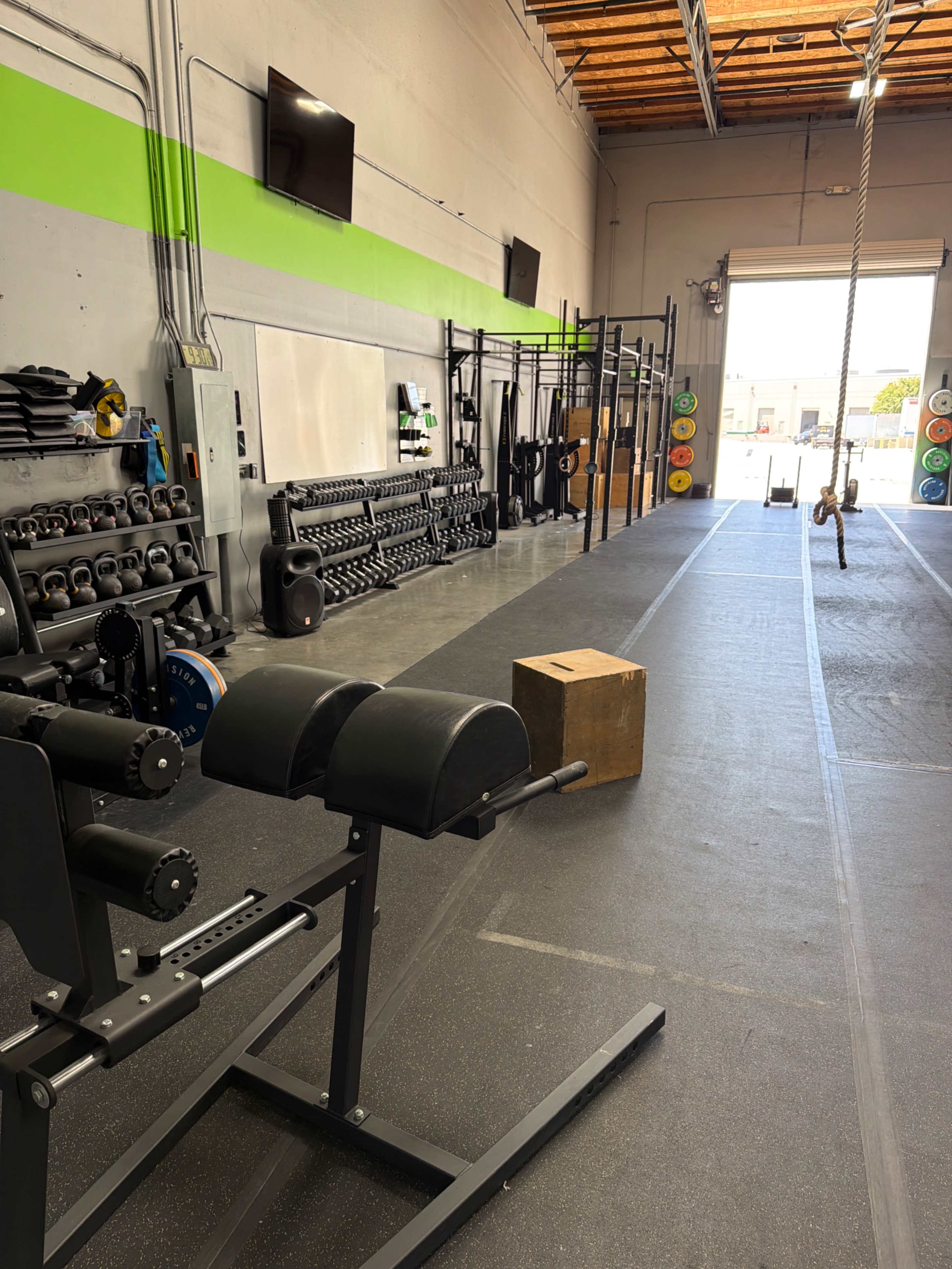 The image depicts a gym interior featuring various weightlifting equipment, a wooden box, and a rope hanging from the ceiling, with bright green accents on the walls.