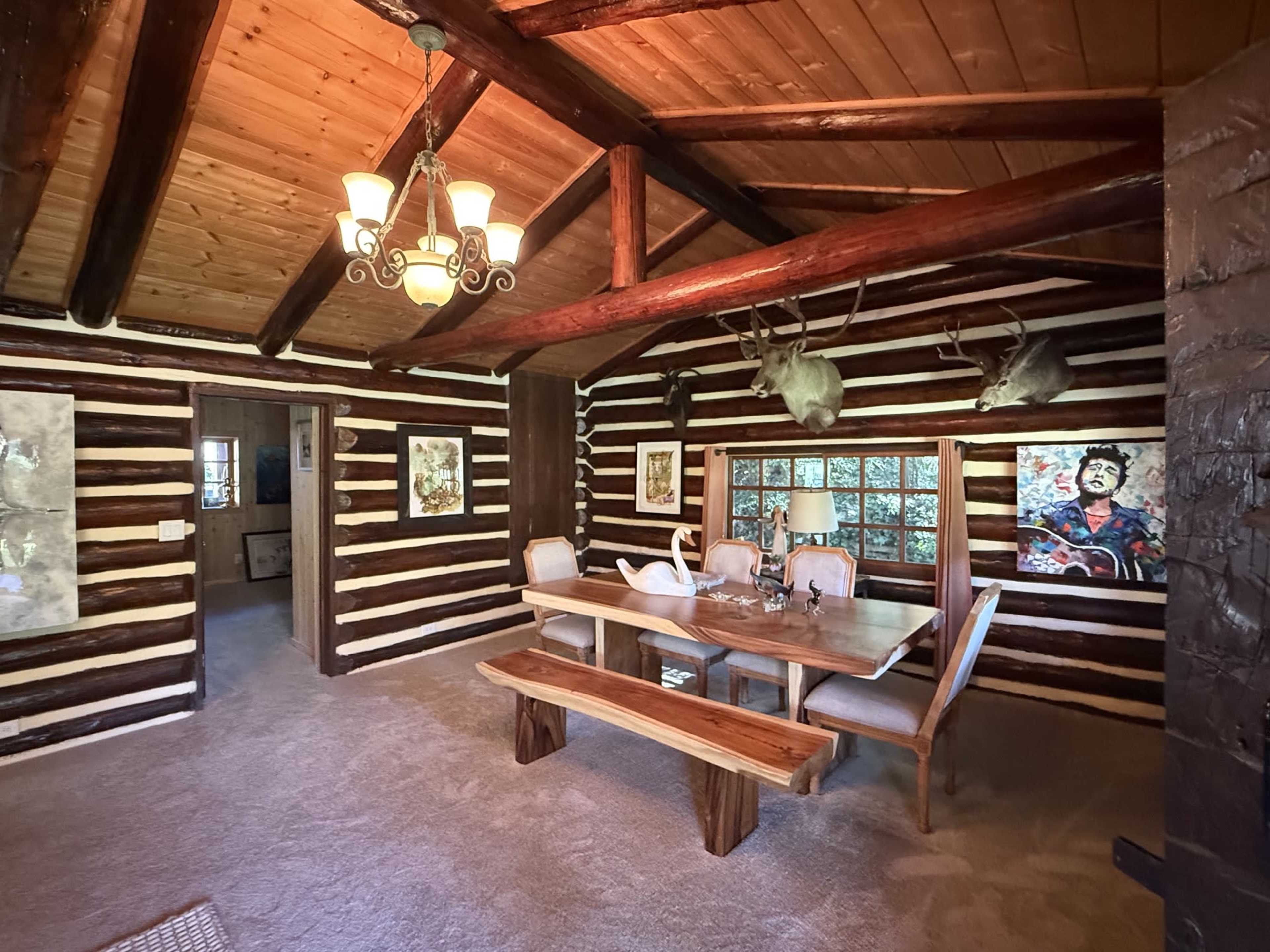 The image shows a rustic dining area with a large wooden table and benches, surrounded by log walls and a ceiling with exposed beams.