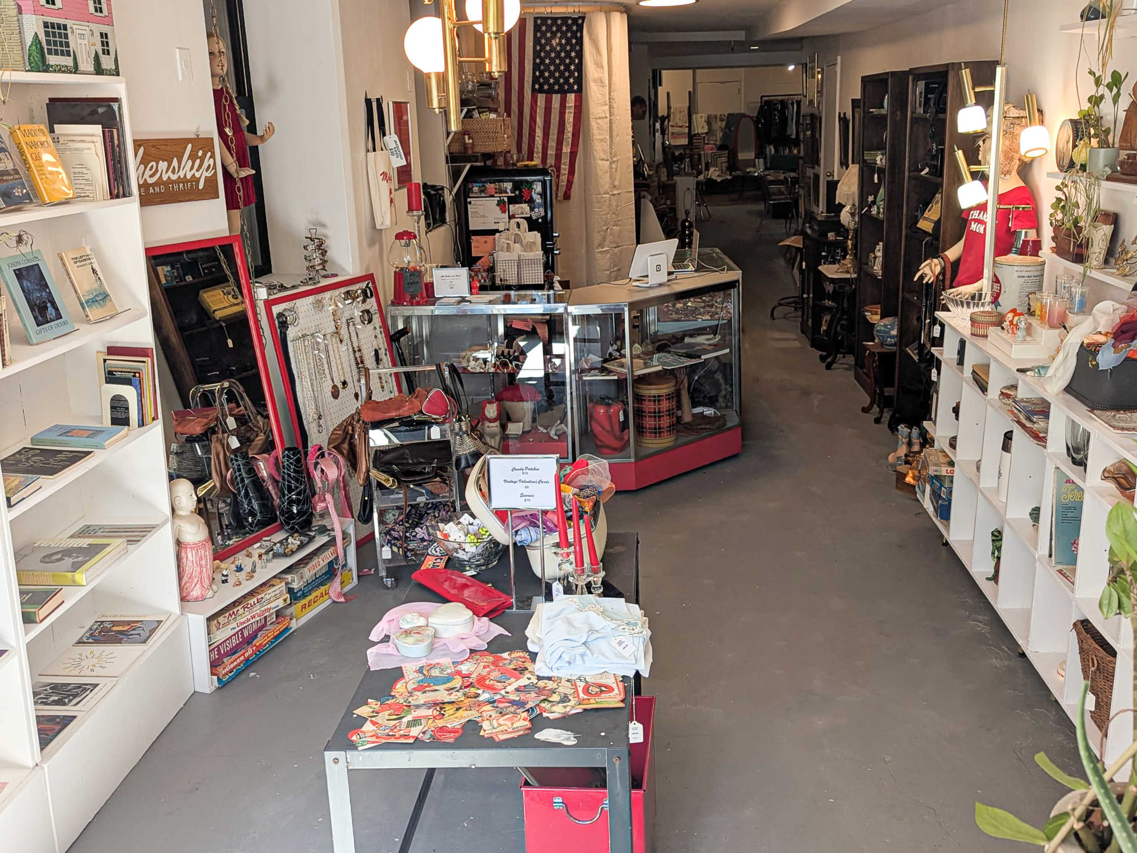 The image shows an interior of a shop displaying various vintage items, books, and clothes, with an American flag hanging on the wall.
