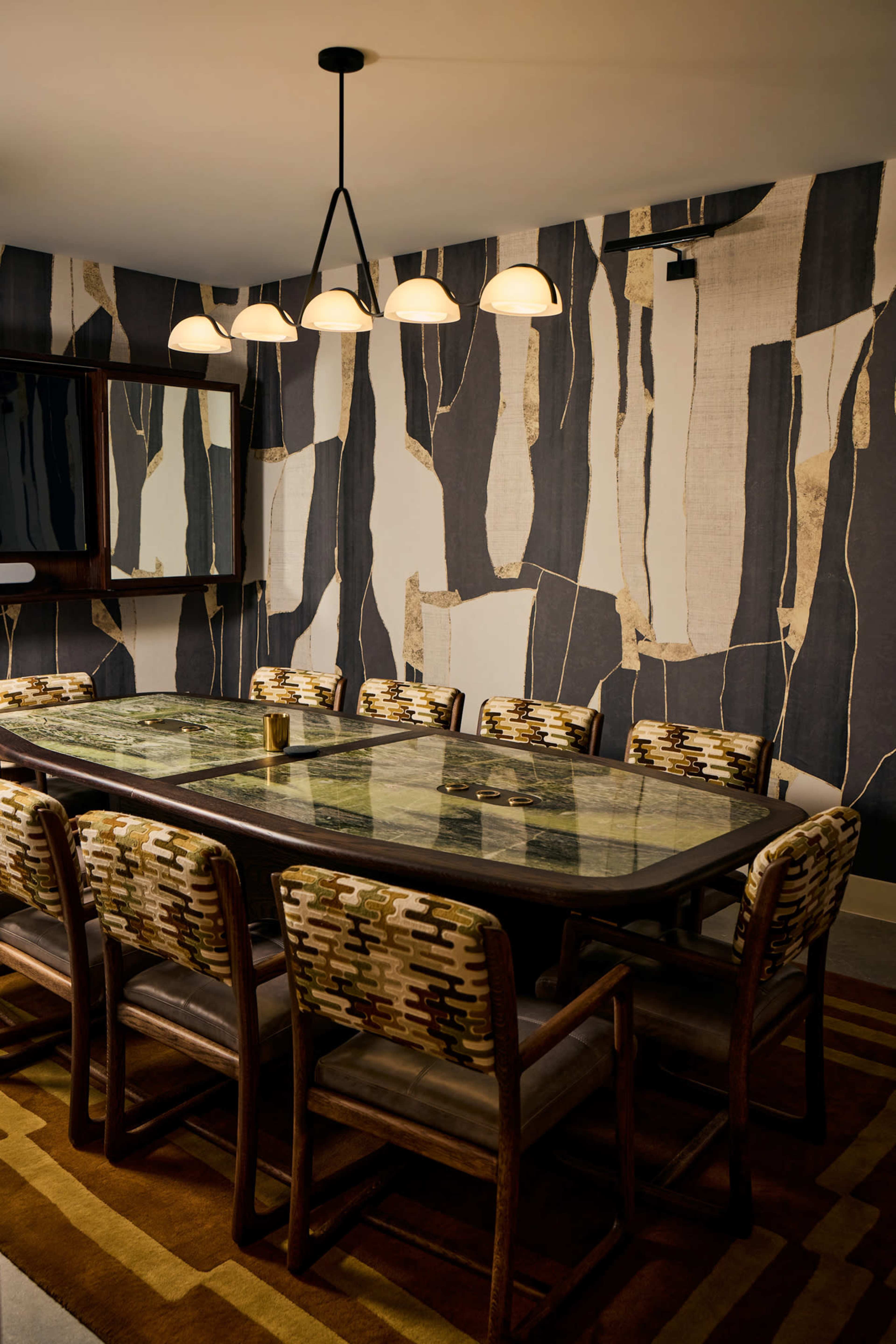 A modern dining room features a large glass-top table surrounded by upholstered chairs, set against a patterned wallpaper with abstract designs.
