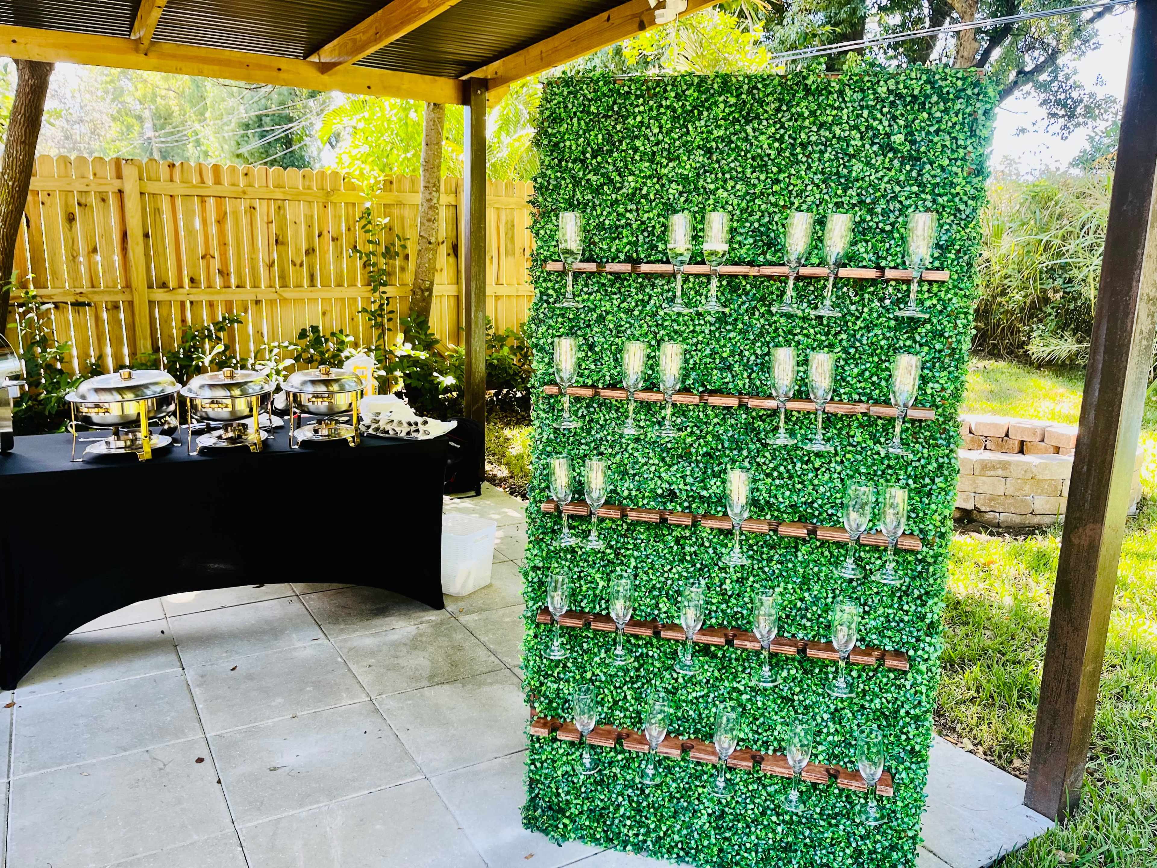 A green wall covered in foliage decorations stands next to a table with food serving trays in an outdoor setting.