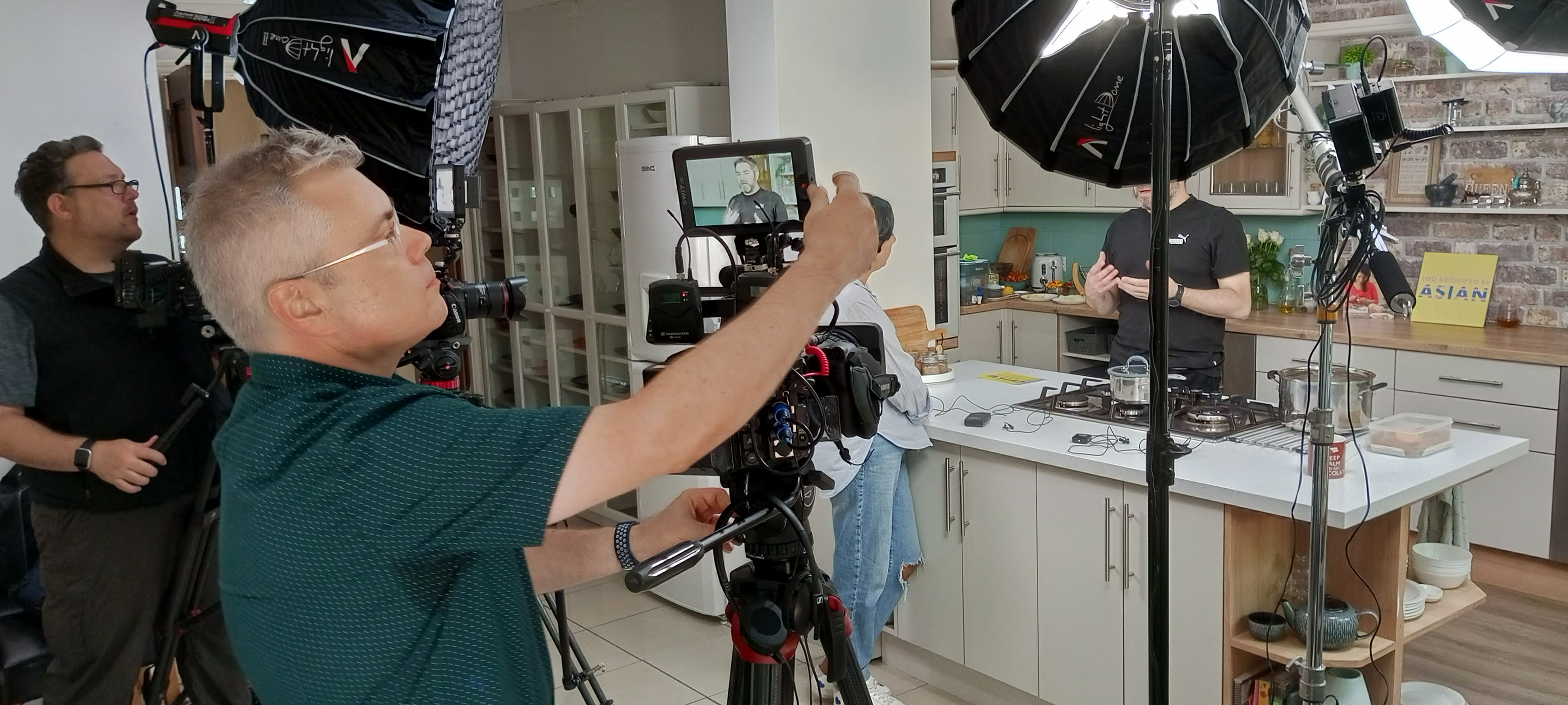 A group of individuals films a cooking demonstration in a kitchen, with cameras and lighting equipment set up to capture the scene.