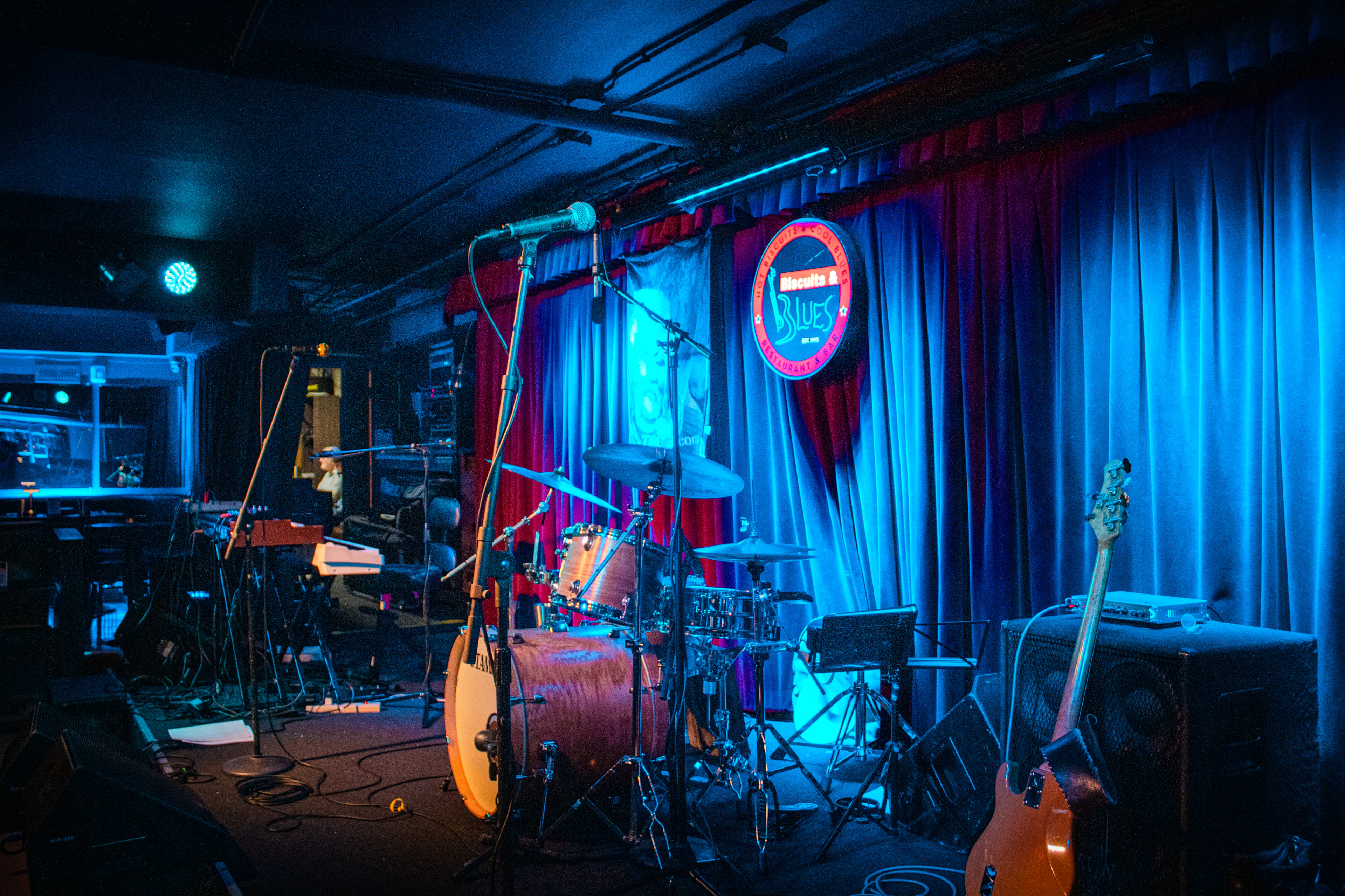 The stage is set with musical instruments, including a drum kit and a bass guitar, in a dimly lit venue adorned with blue and red curtains.