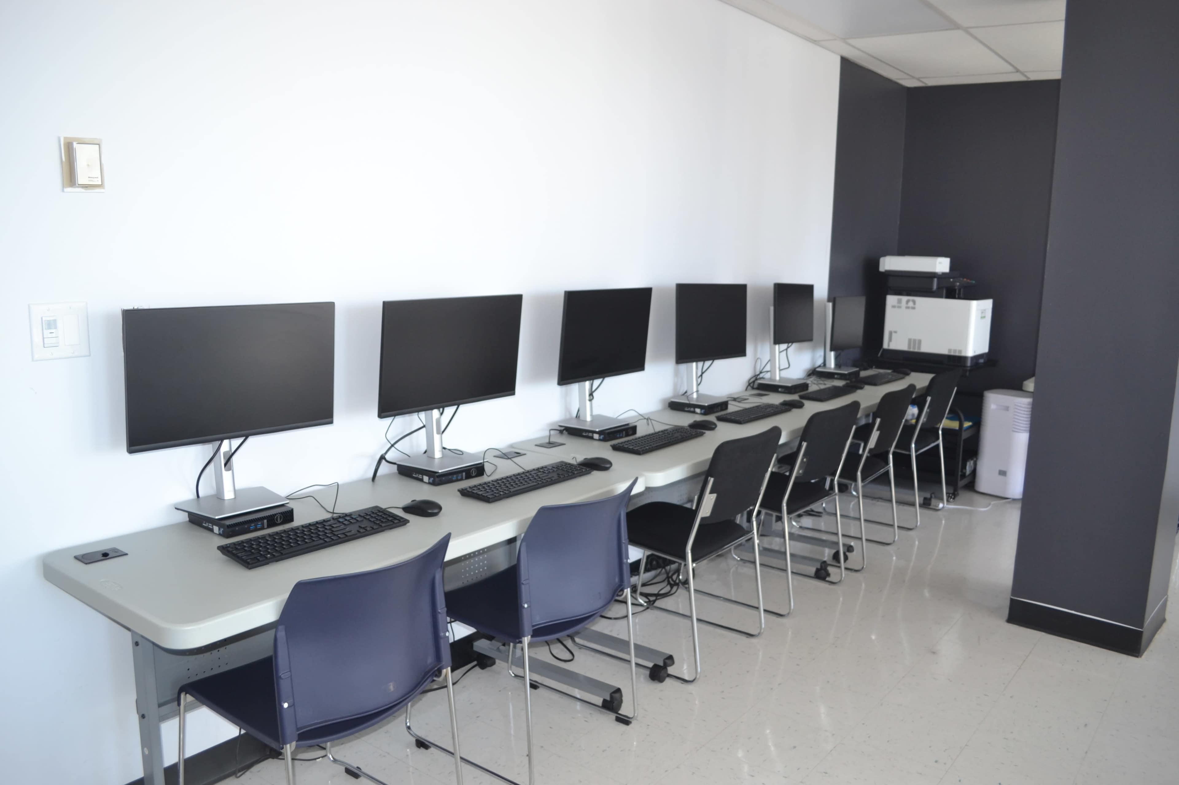 A row of computer workstations with monitors, keyboards, and chairs is set against a plain wall in a well-lit room.