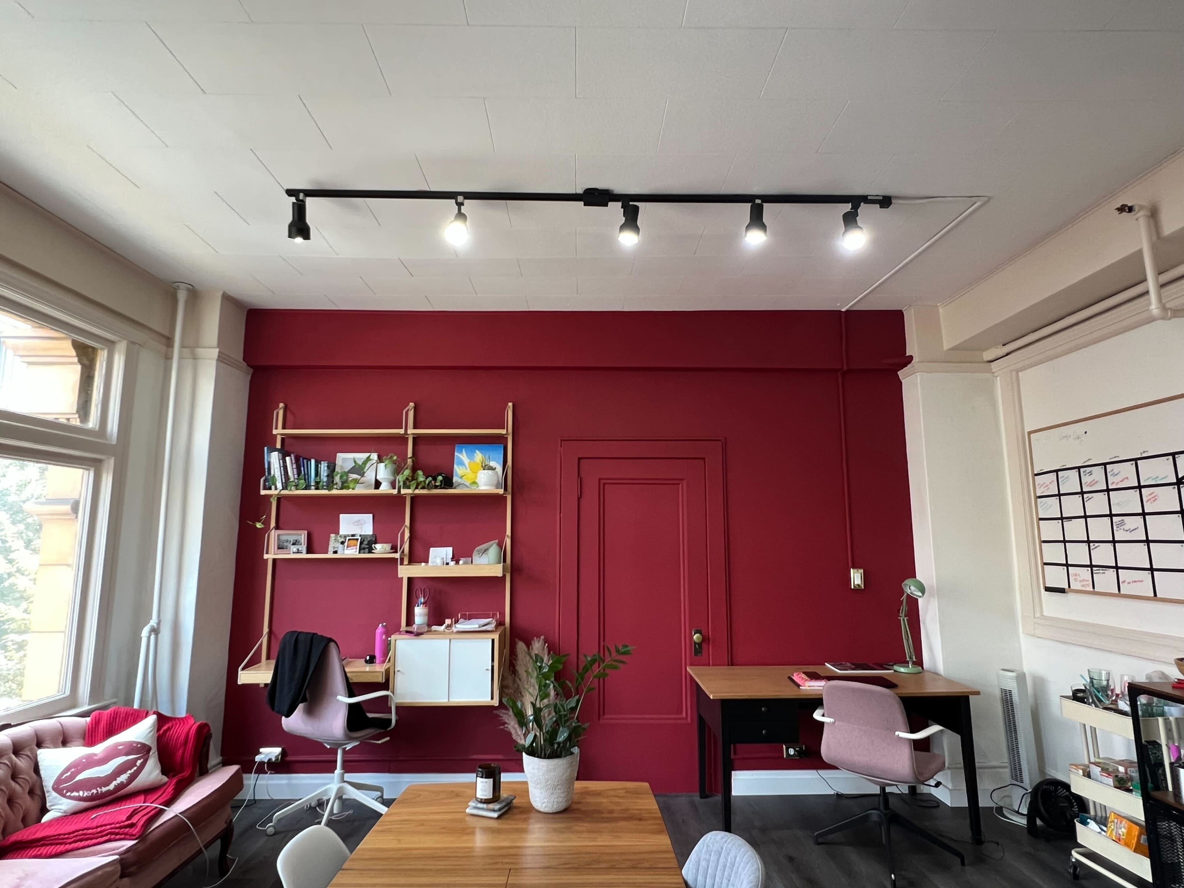 A modern office space with a maroon wall, wooden table, pink chairs, and a shelf filled with books and decor.