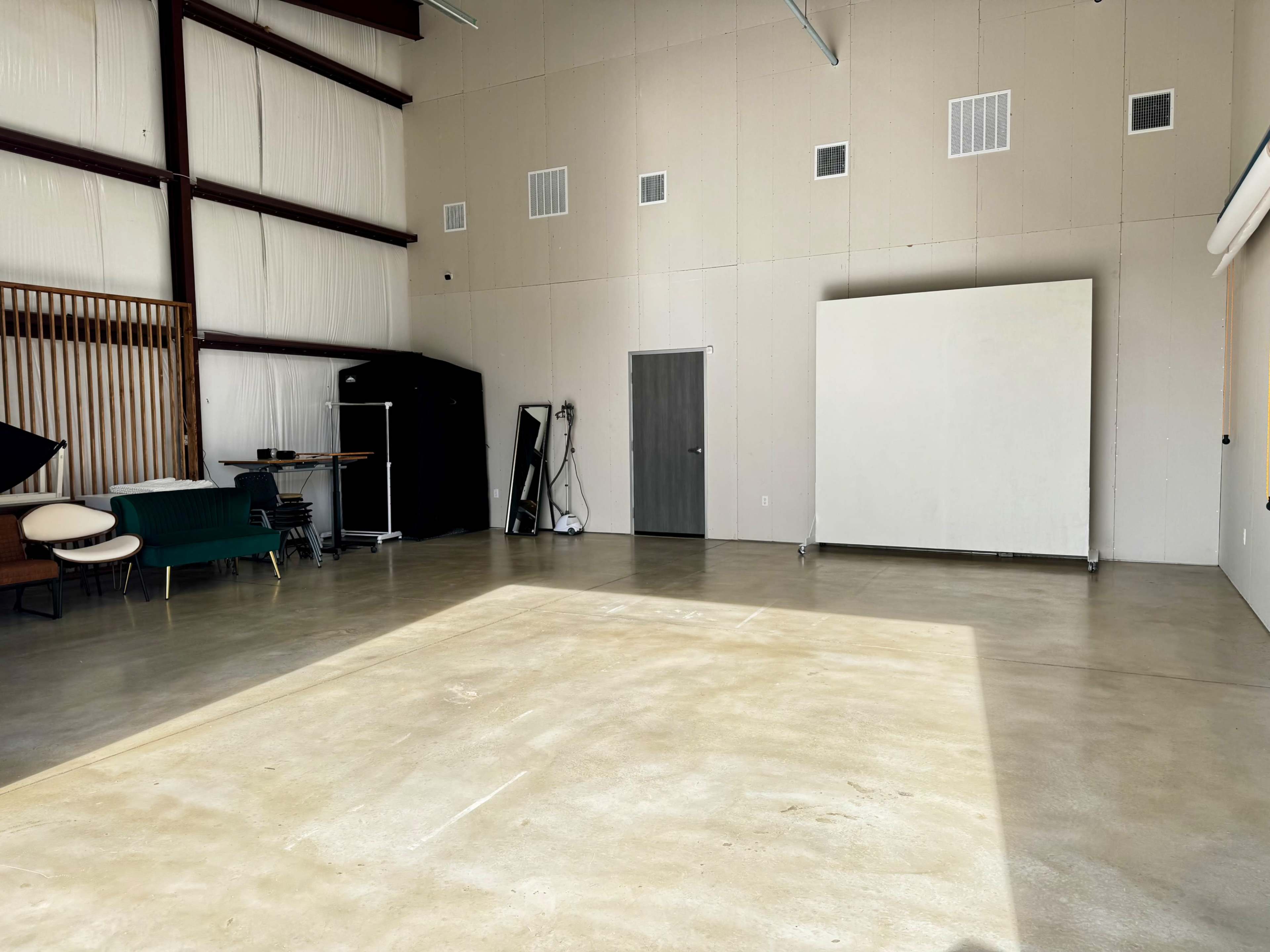 The image shows a spacious, mostly empty industrial room with concrete flooring and high metal ceilings, featuring a few pieces of furniture and a large white wall.