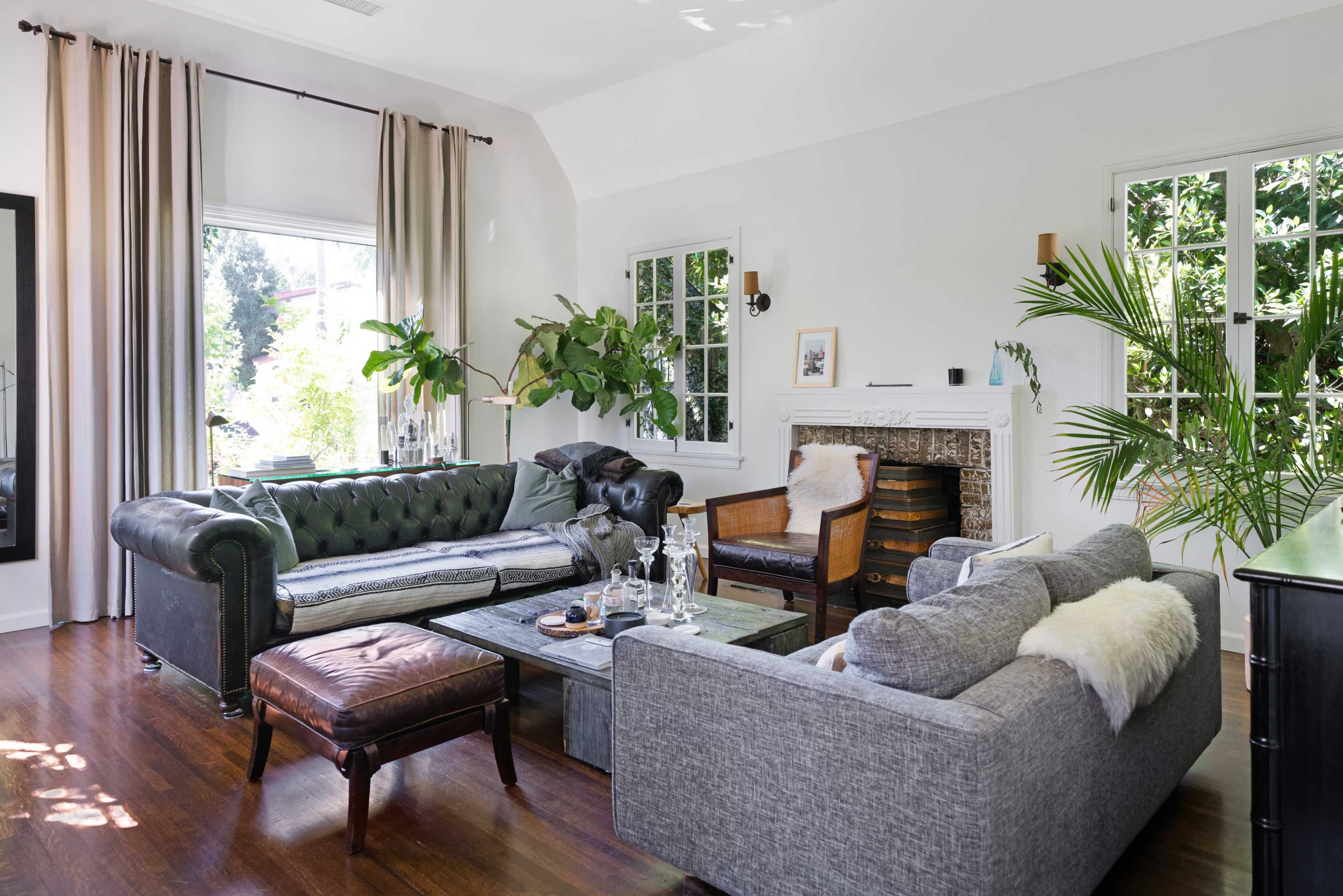 The living room features a mix of a dark green chesterfield sofa, a gray sectional, and a wooden armchair, arranged around a coffee table, with abundant natural light from large windows.