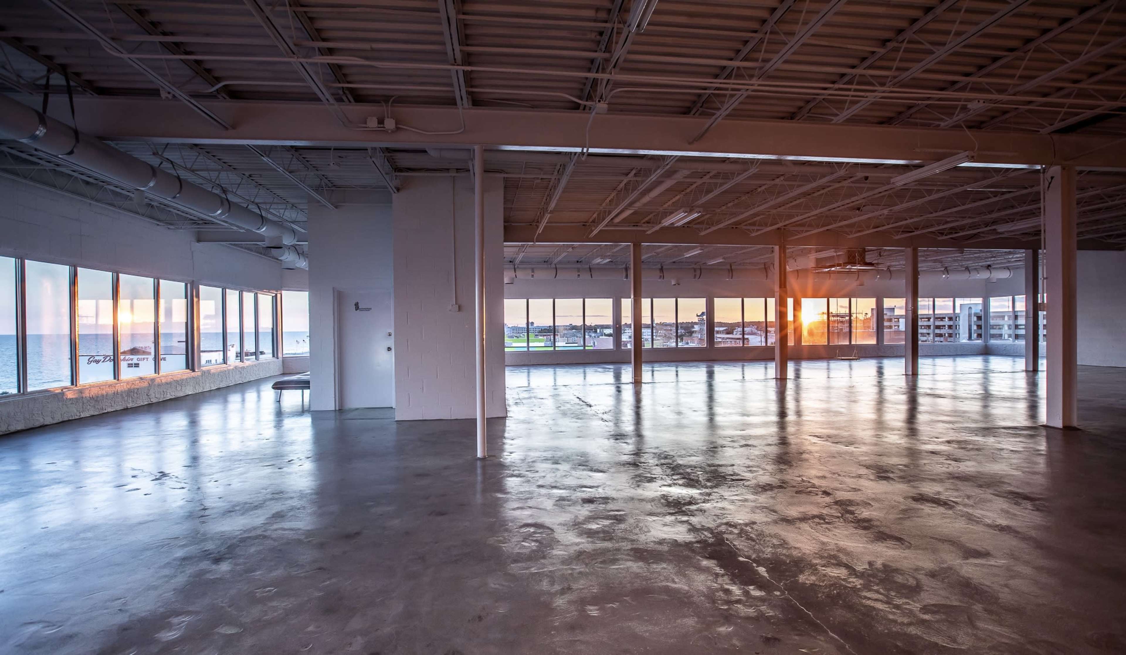 The image shows an empty, spacious interior with large windows displaying a view of a waterfront at sunset.