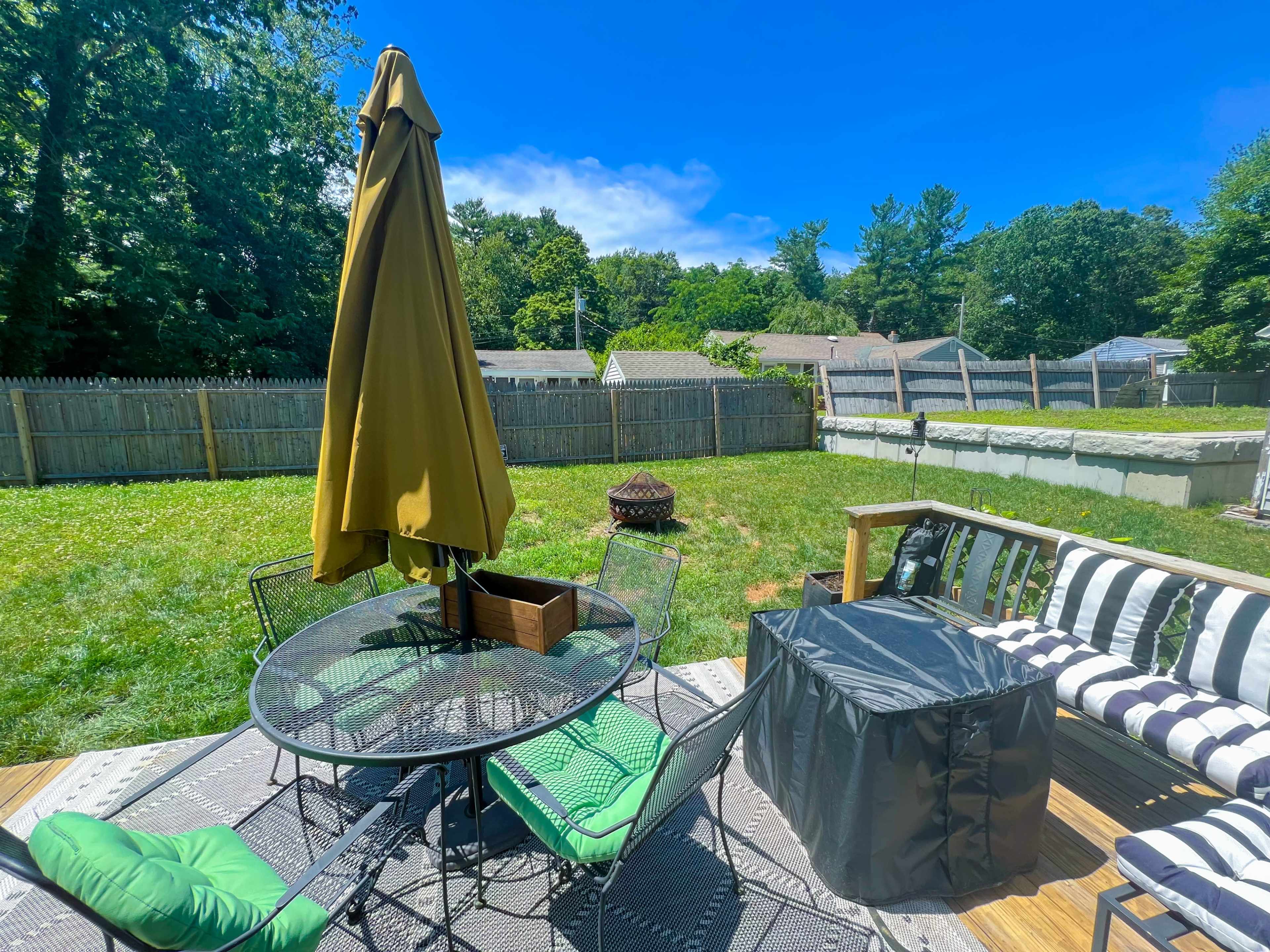 A backyard patio with a round table and chairs, a large umbrella, and a seating area, all set on a wooden deck with a grassy lawn behind.