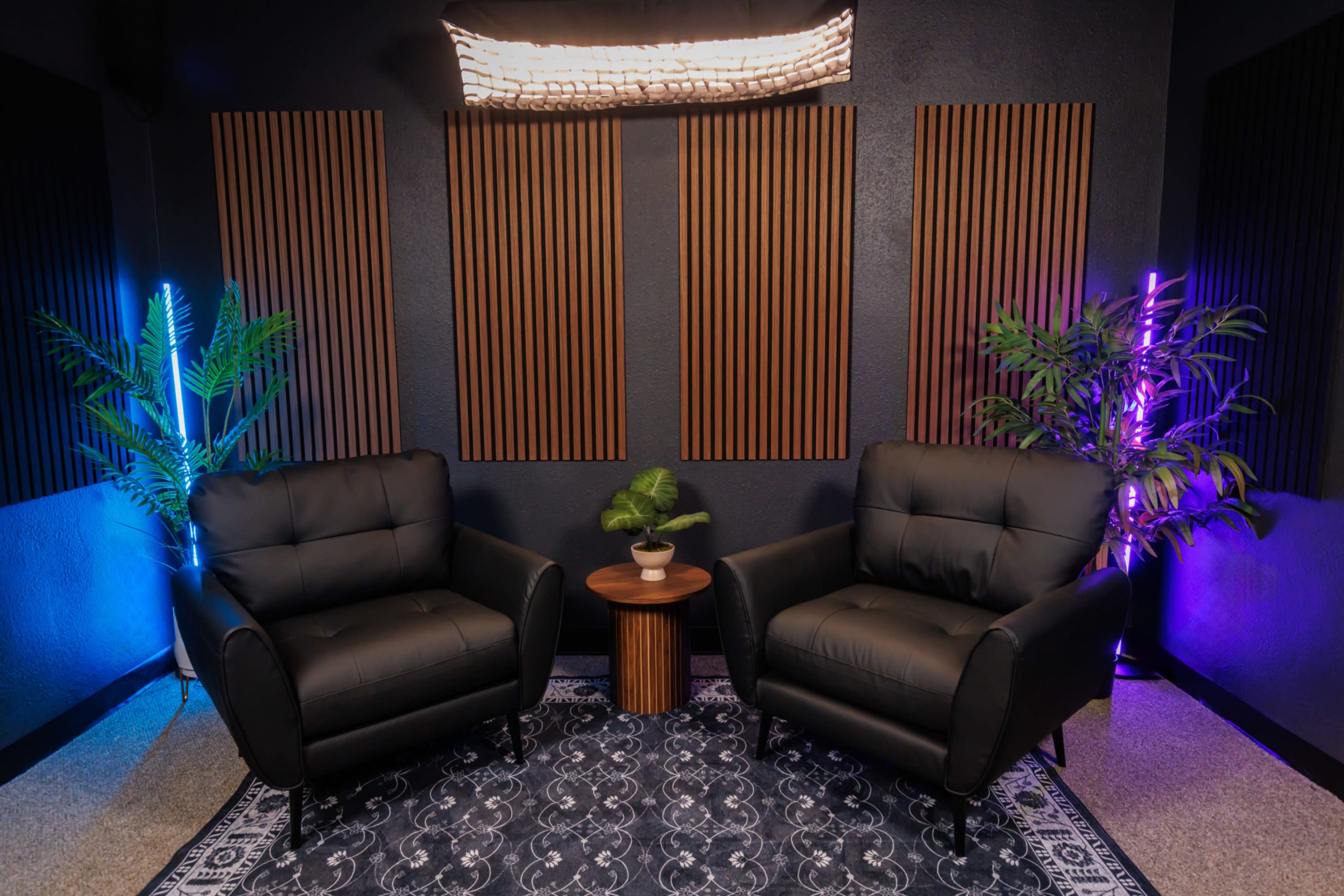 The image shows a cozy seating area with two black armchairs flanking a small wooden table, surrounded by decorative plants and ambient lighting in a room with wooden wall panels.