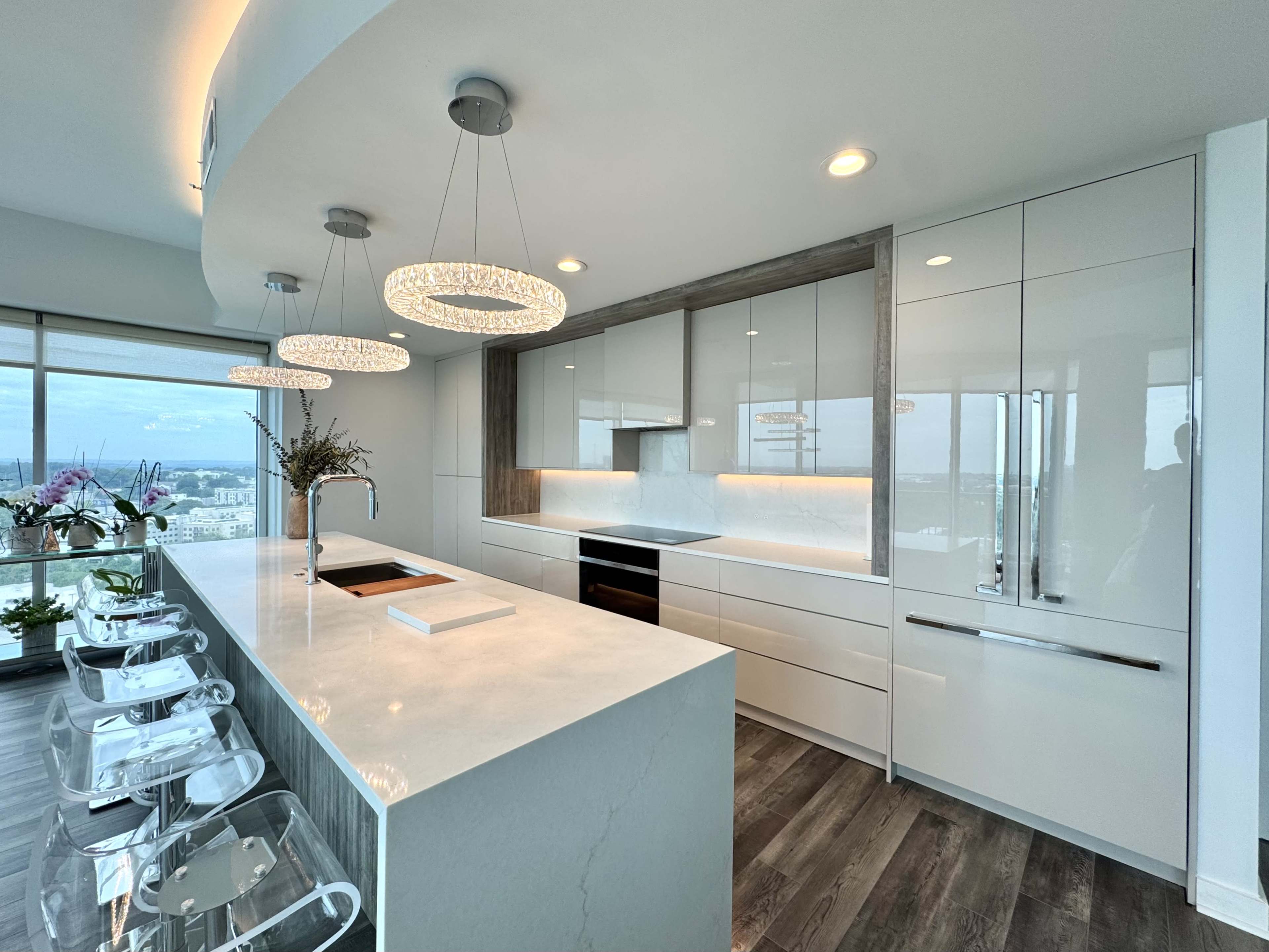 The image shows a modern kitchen with sleek white cabinets, a large island with a marble countertop, and pendant lighting above.