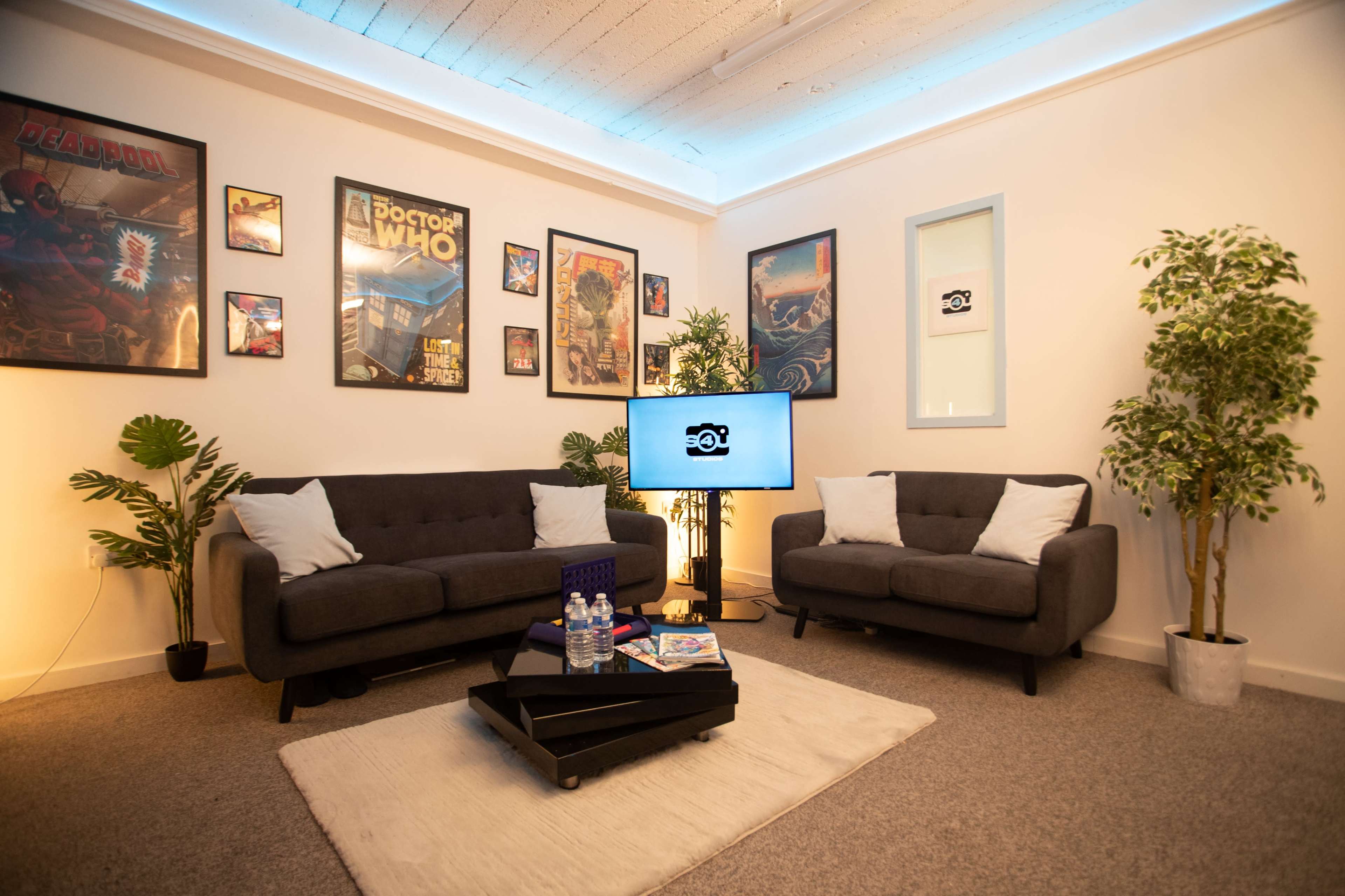 A modern lounge area with two gray sofas, framed posters on the walls, a television on a stand, and a coffee table with books and water bottles.