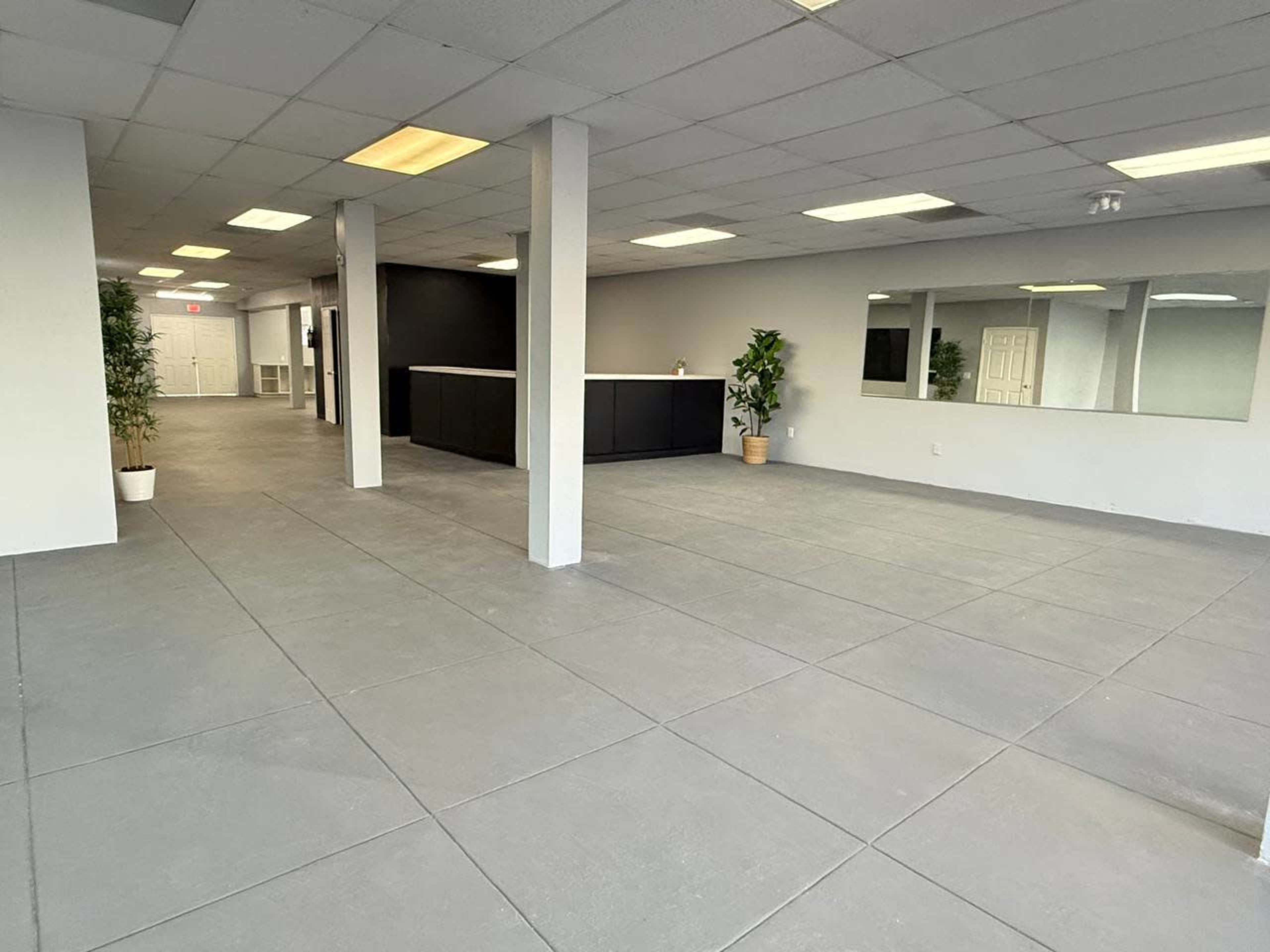 The image shows a spacious, empty commercial interior with gray flooring, several lights, and minimal furnishings, including potted plants and a reception area in the background.