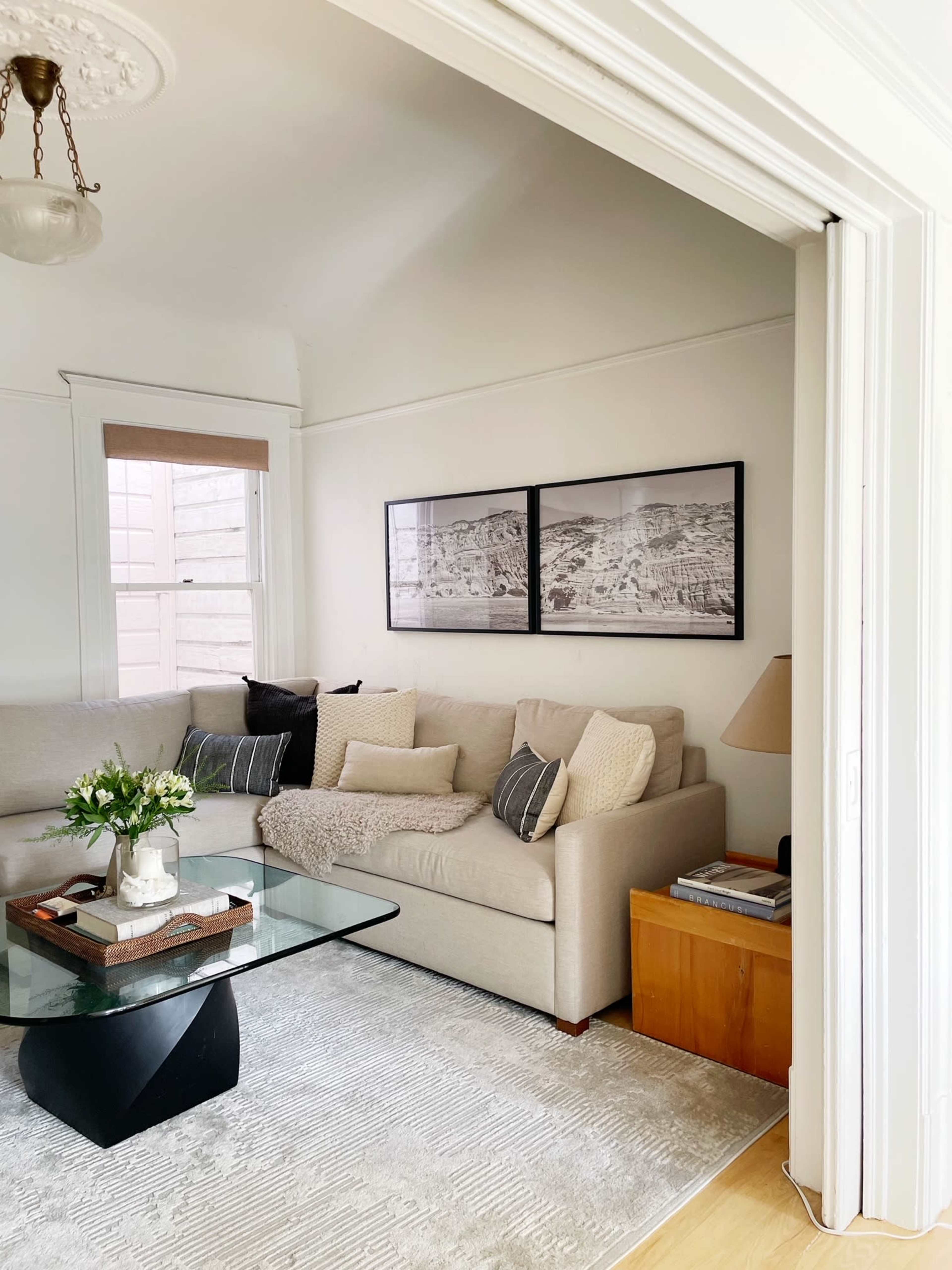 A living room with a light-colored sectional sofa, a glass coffee table, framed black-and-white photographs on the wall, and a lamp next to a wooden side table.