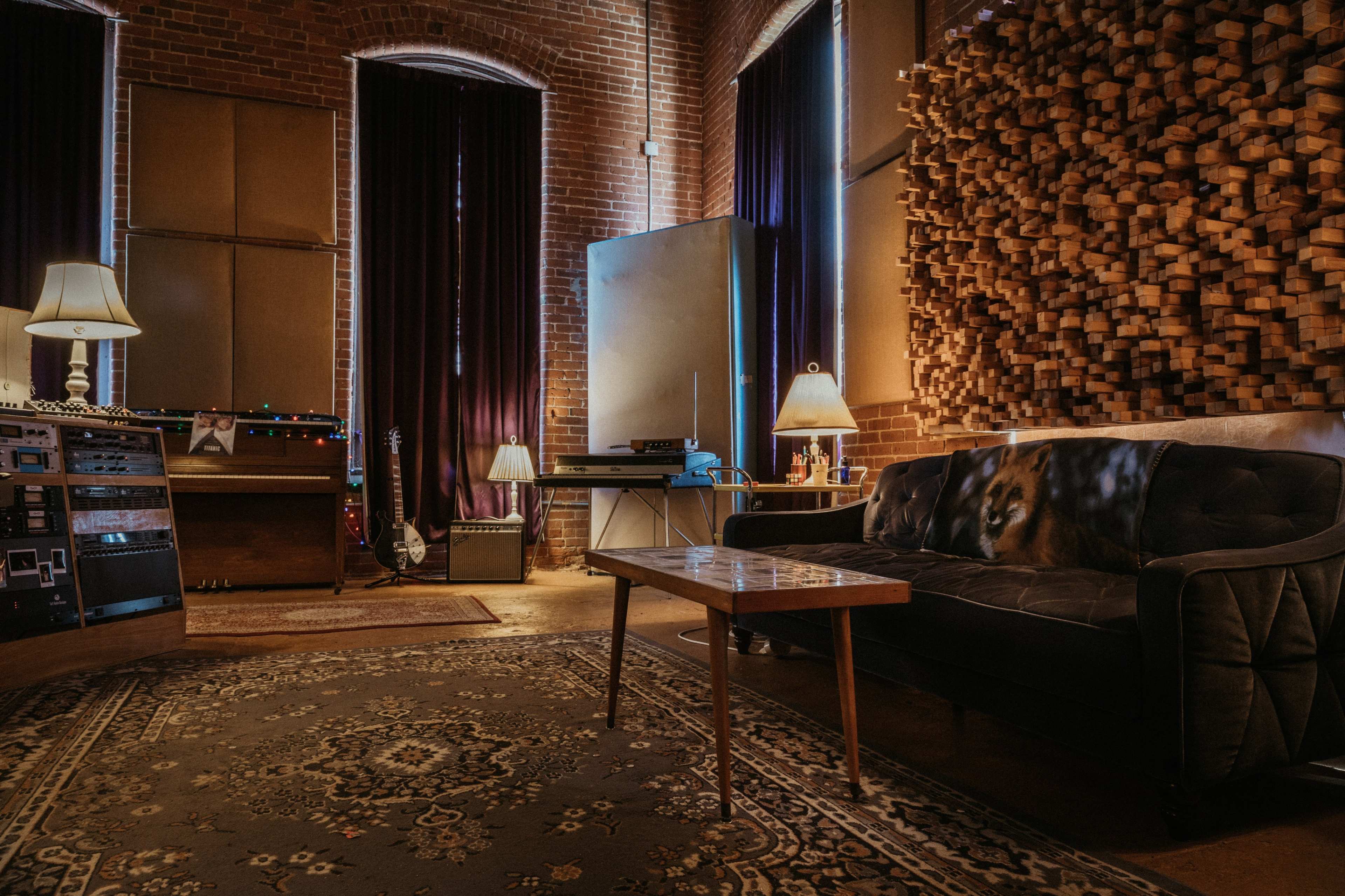 A cozy music studio features a black couch, wooden sound paneling, a vintage recording console, and soft lighting from two lamps.