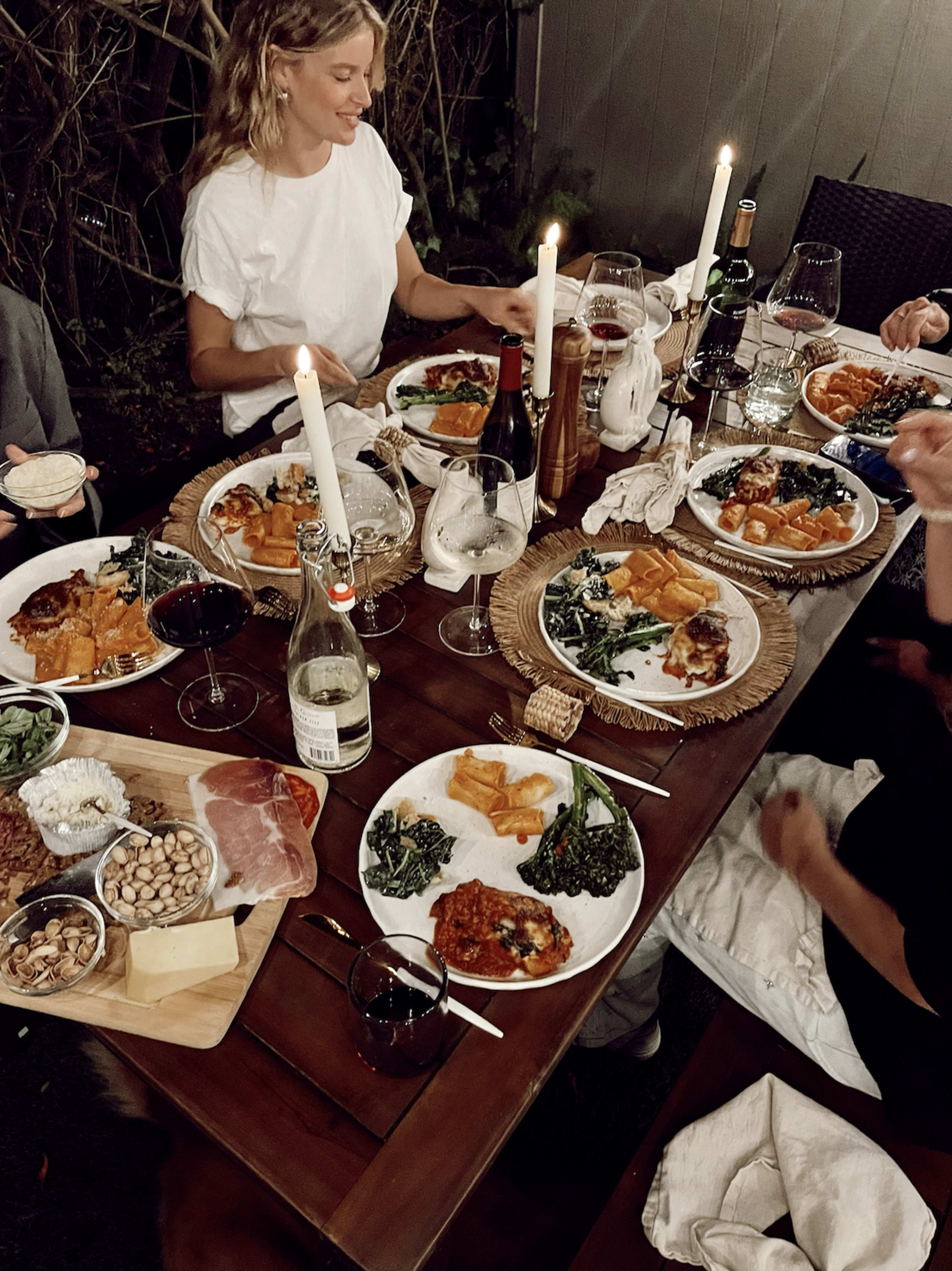 A group of people enjoys a dinner spread featuring various dishes, wine, and candles on a wooden table outdoors.