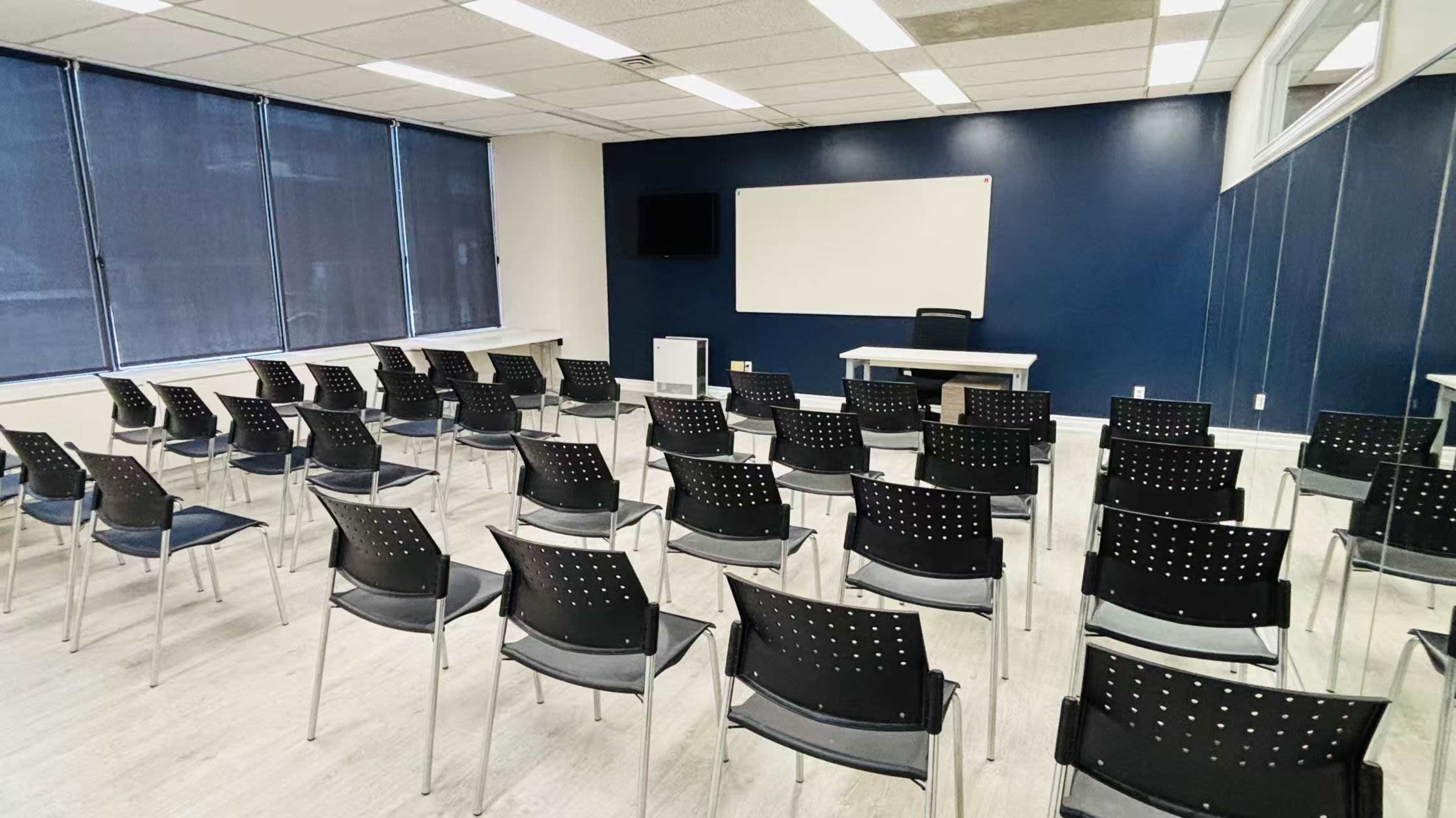 The image shows a classroom setting with neatly arranged black chairs facing a blank whiteboard and a television on a blue accent wall.