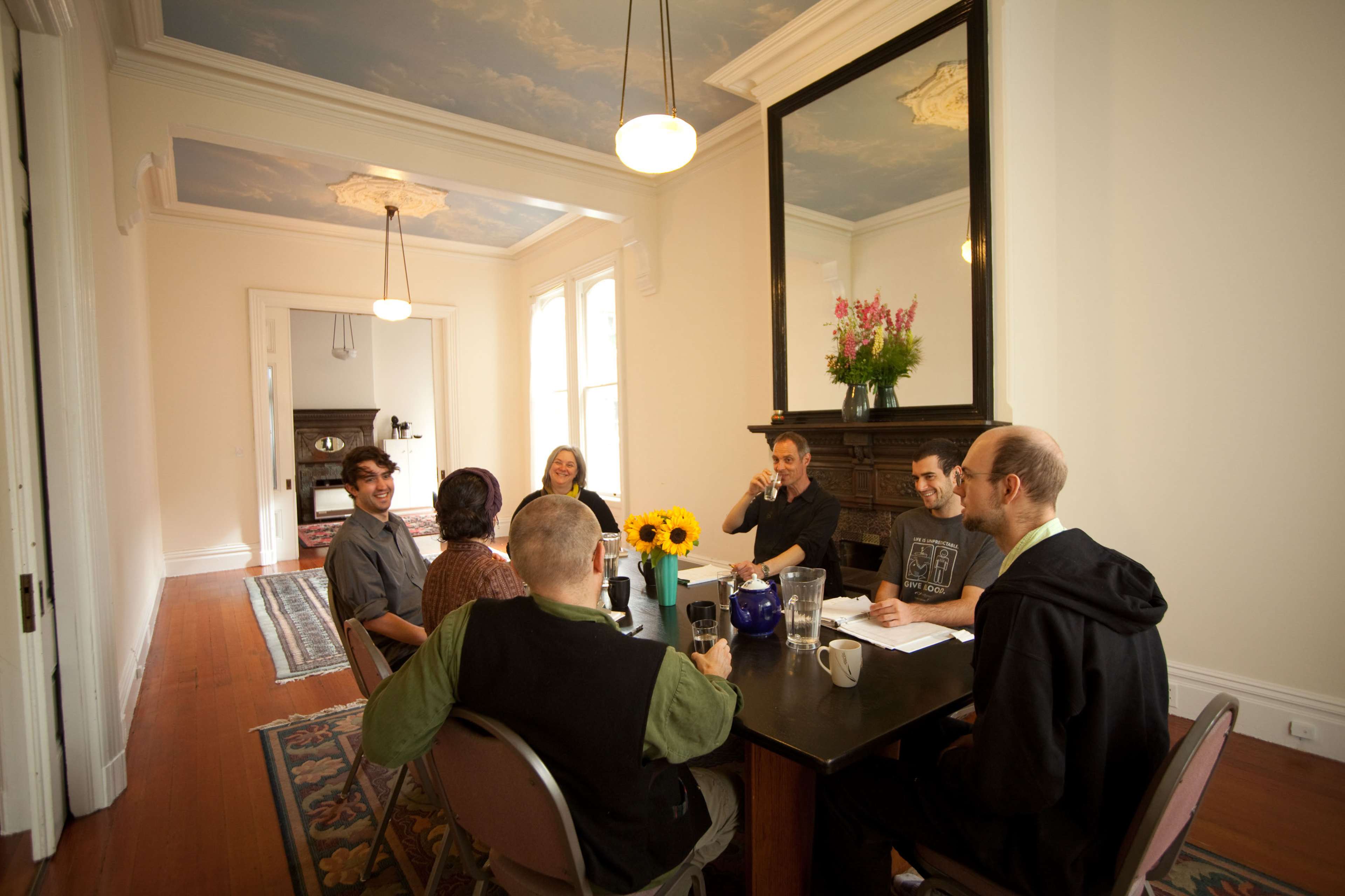 A group of six people is seated around a dining table in a spacious room with a mirror and a floral arrangement.