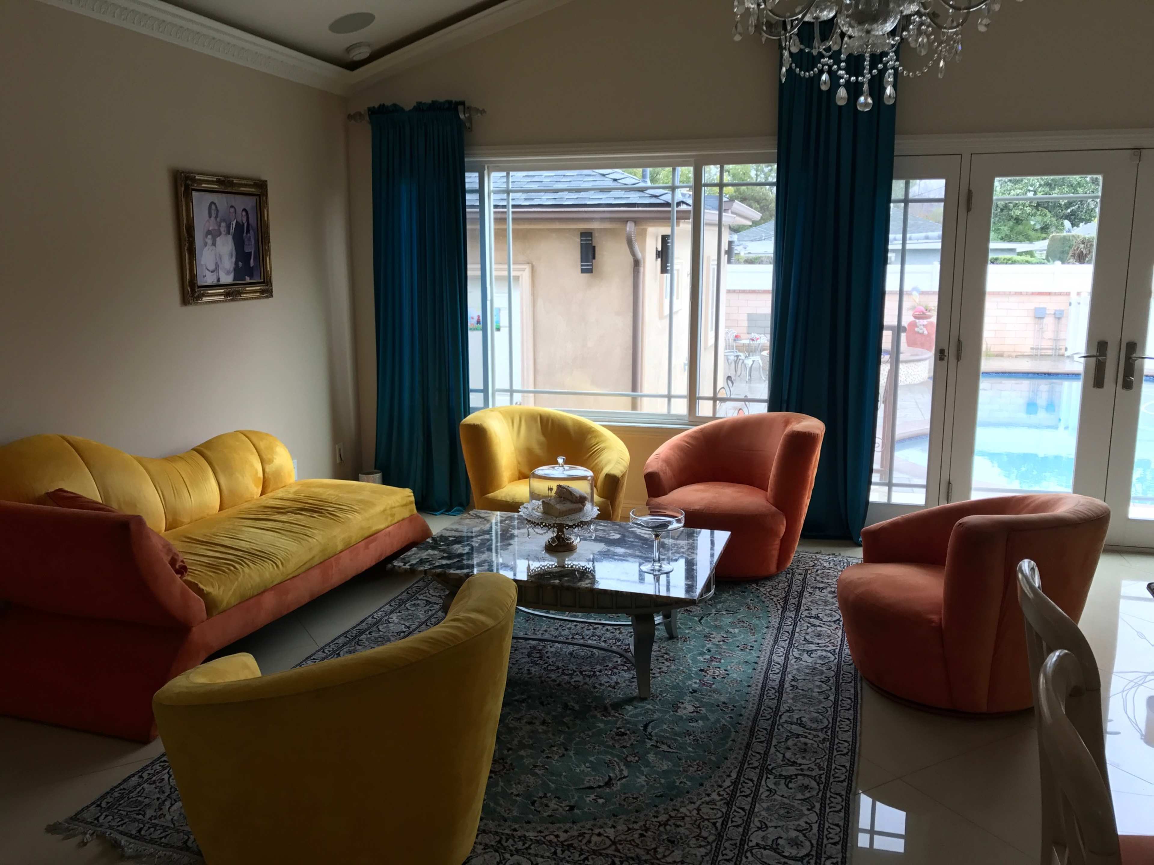 A brightly colored seating area features large, rounded chairs and a sofa arranged around a glass coffee table, with large windows overlooking a pool area.