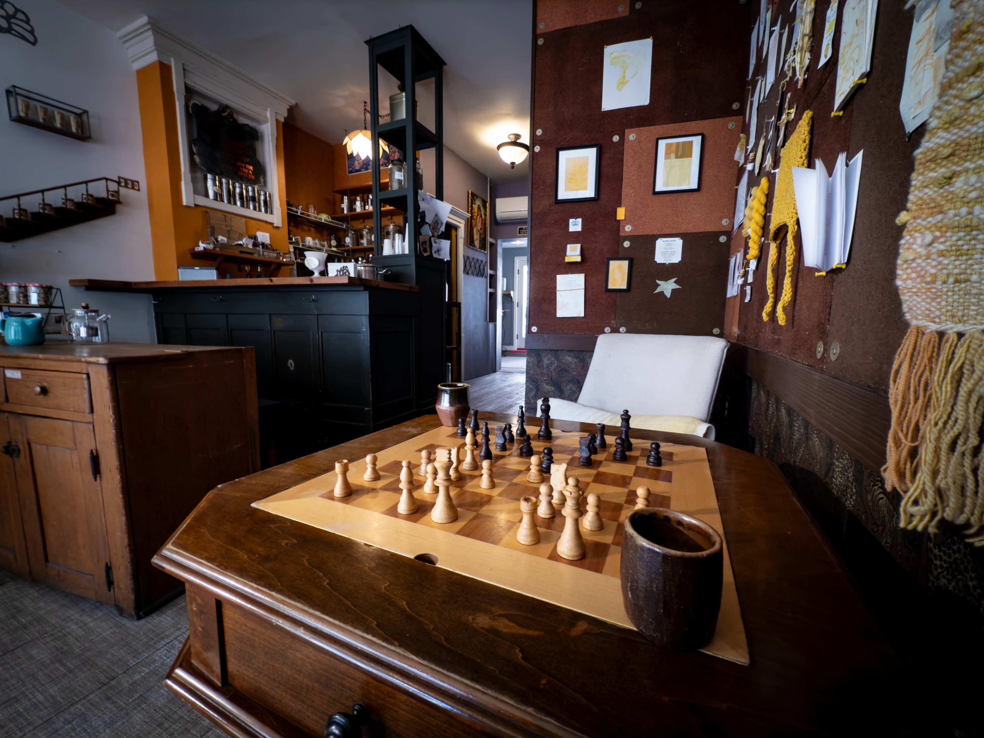A wooden chessboard is set up with pieces arranged on a table in a cozy café with walls adorned with art and notes.
