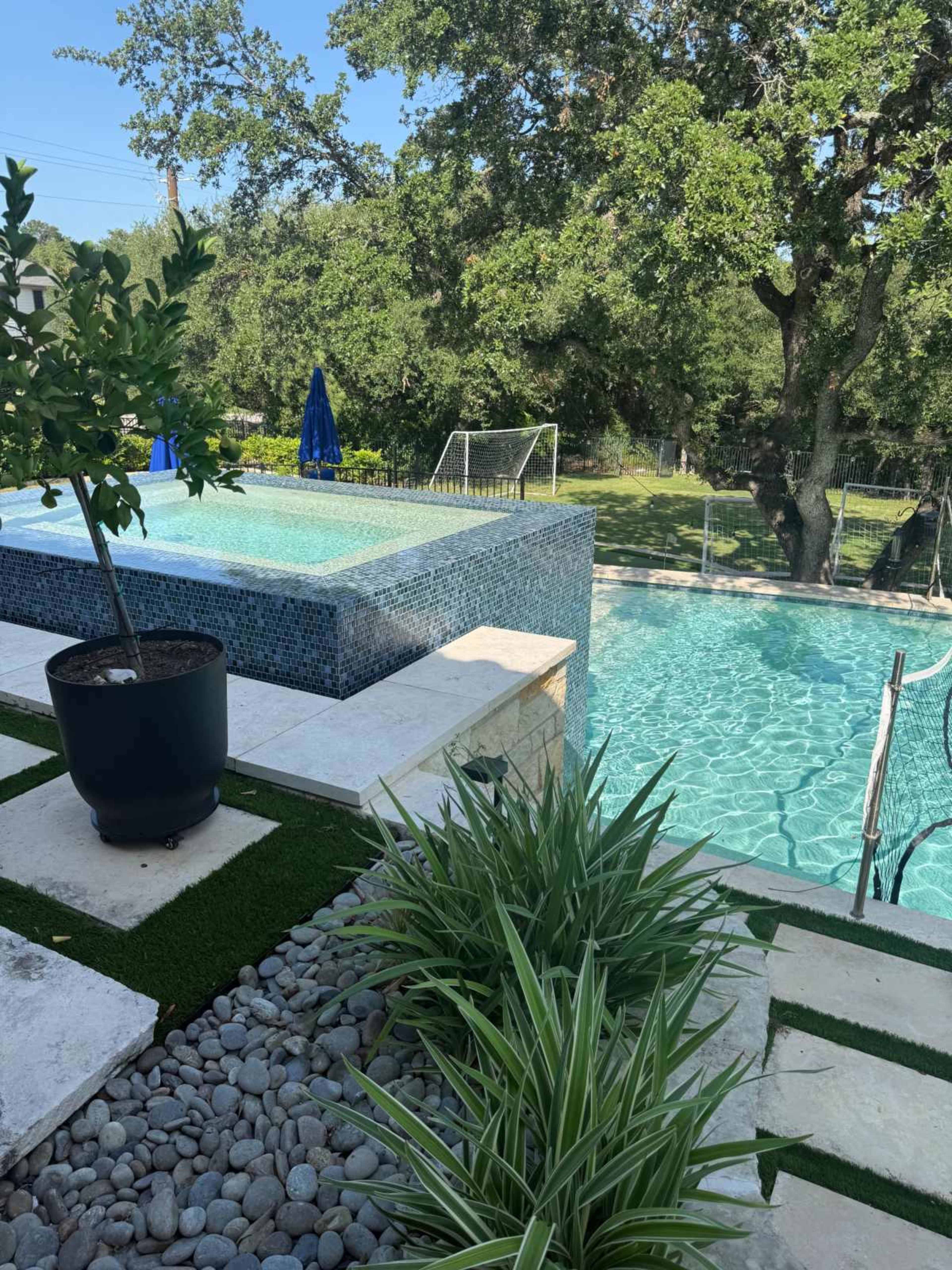 The image shows a landscaped backyard featuring a pool area with a spa, surrounded by greenery and decorative stones.