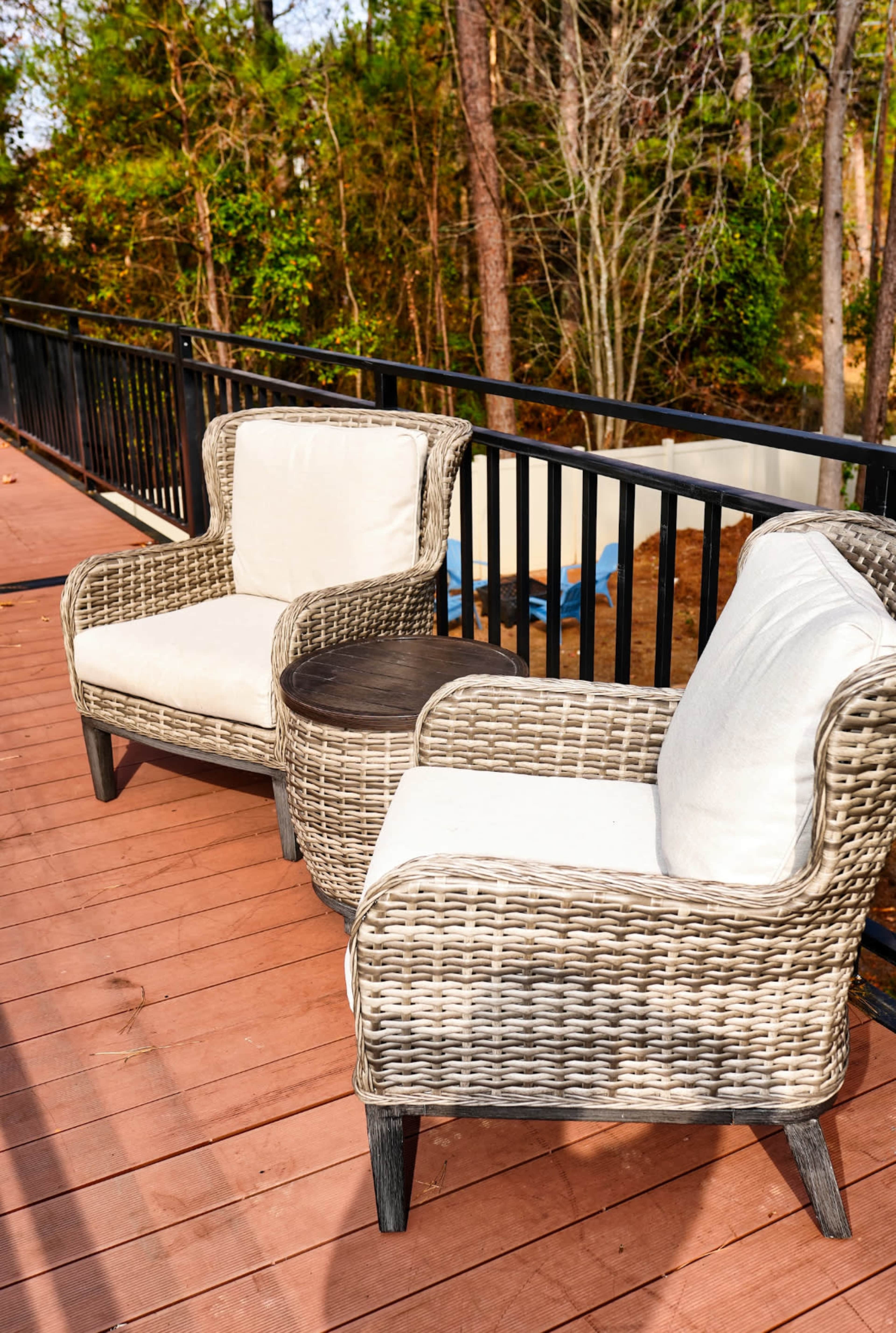 Two wicker chairs with cushions sit beside a small wooden table on a wooden deck overlooking a forested area.