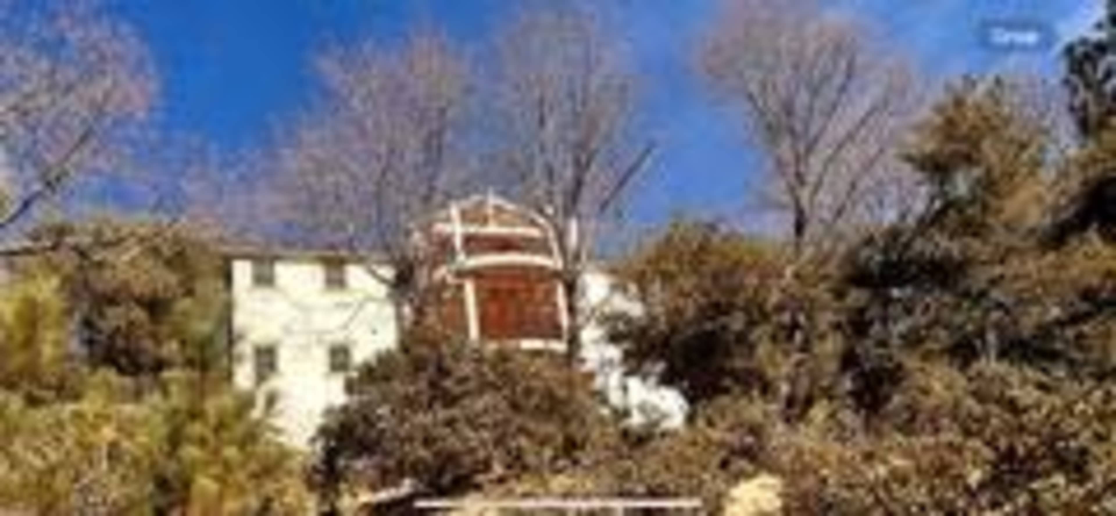 A large, multi-story building with a wooden structure on top is surrounded by trees and shrubs under a blue sky.