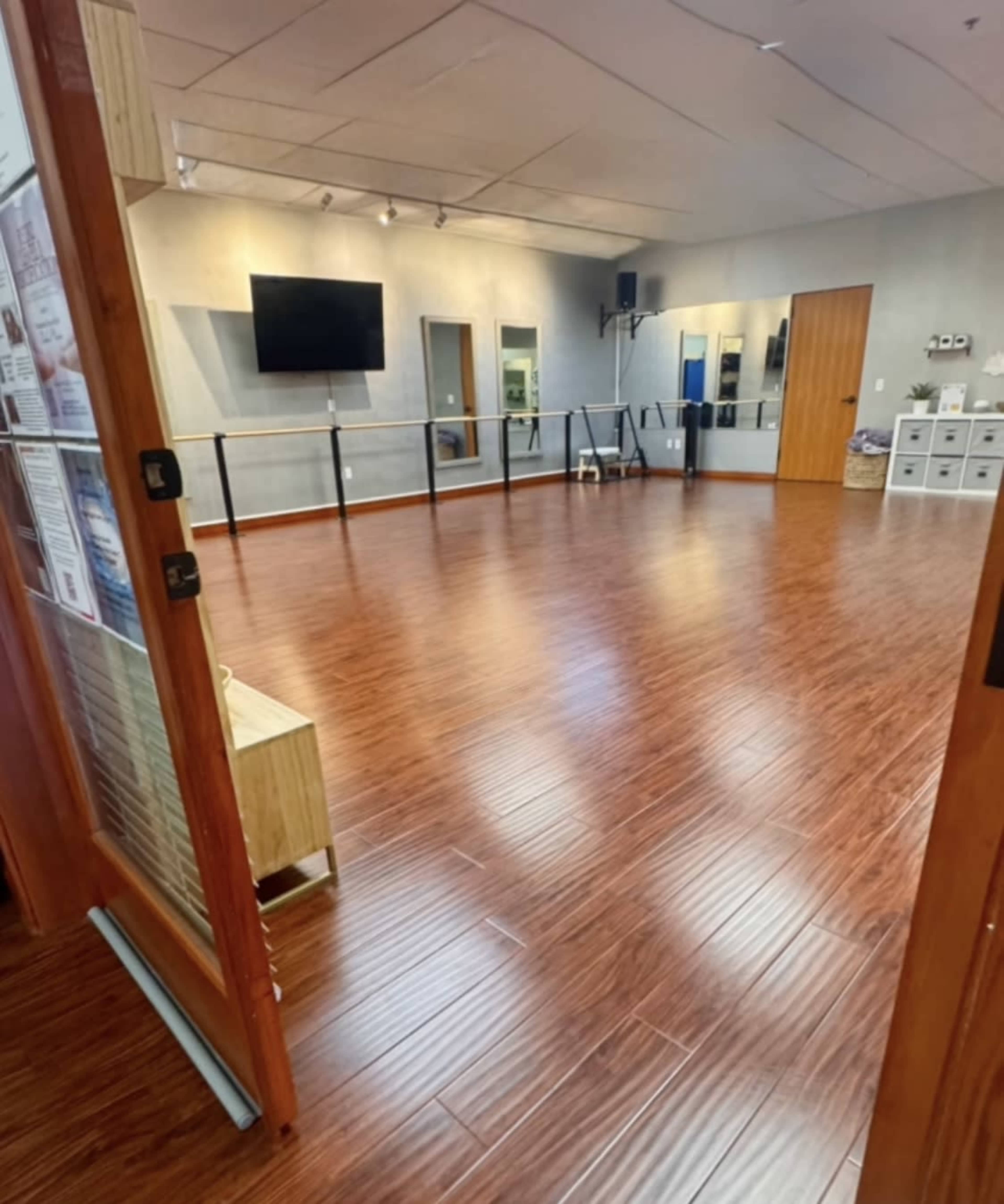 The image shows a spacious dance studio with wooden flooring, a TV mounted on the wall, and mirrors along one side.