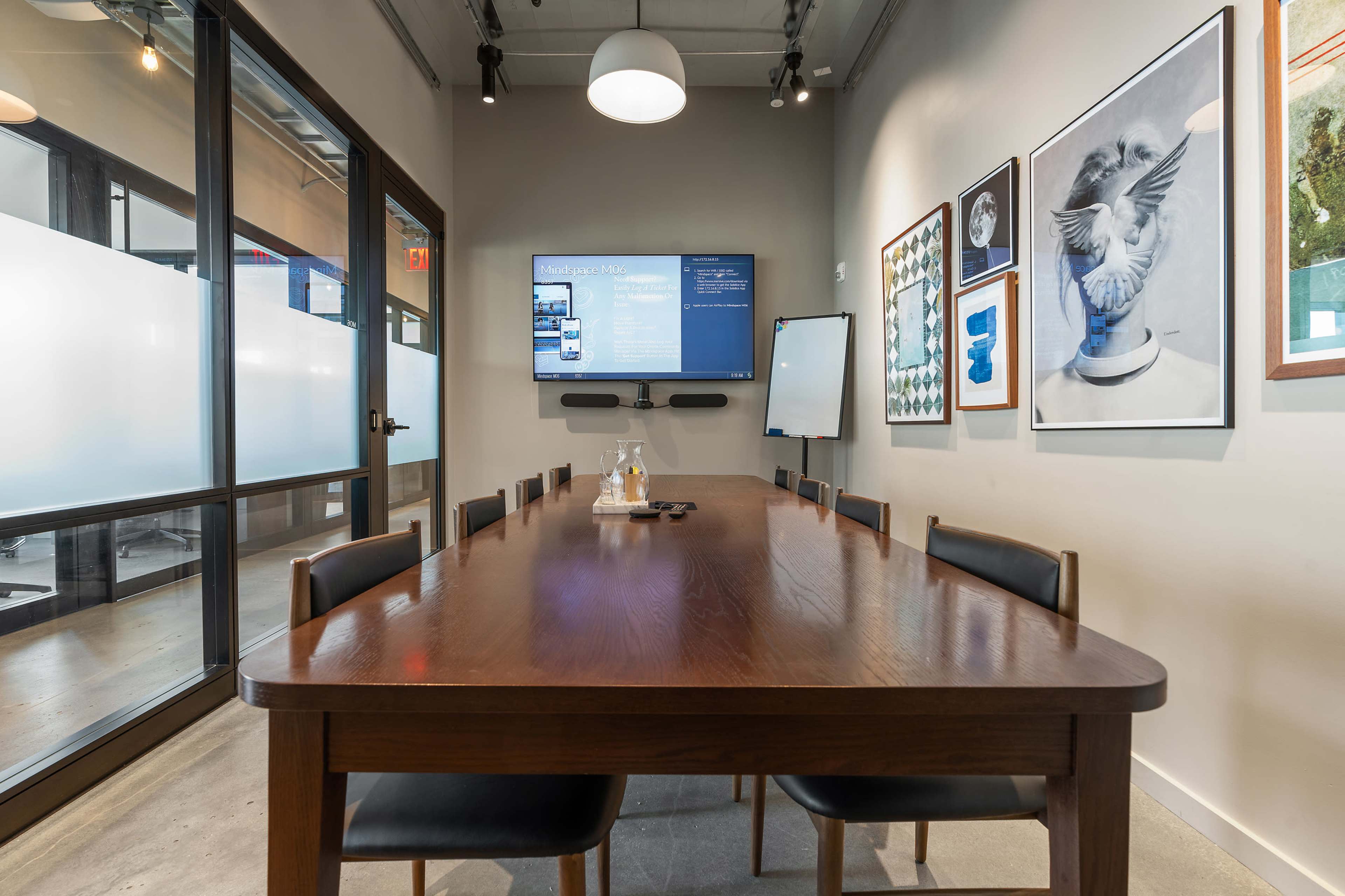 A large wooden conference table is set in a well-lit meeting room, featuring a television screen on the wall and several framed artworks.