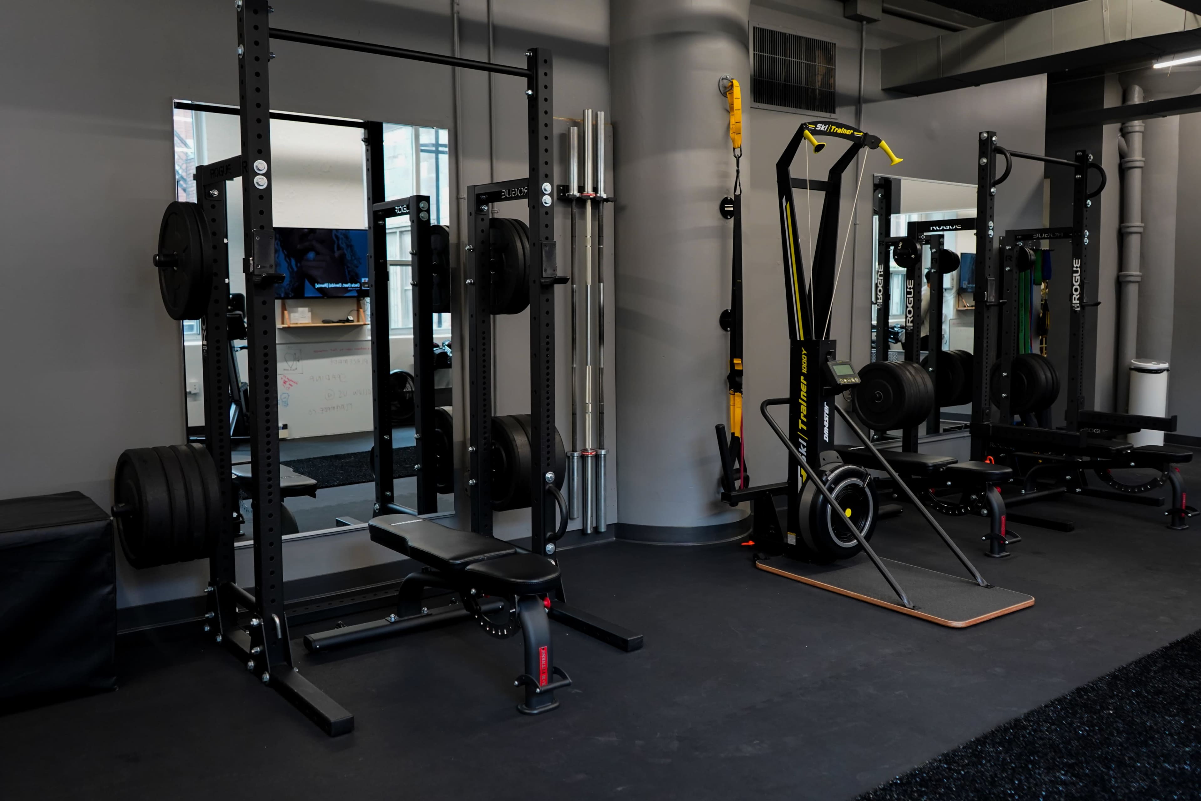 A gym interior with weightlifting equipment, including a squat rack and a bench, alongside a cable machine and a functional trainer.